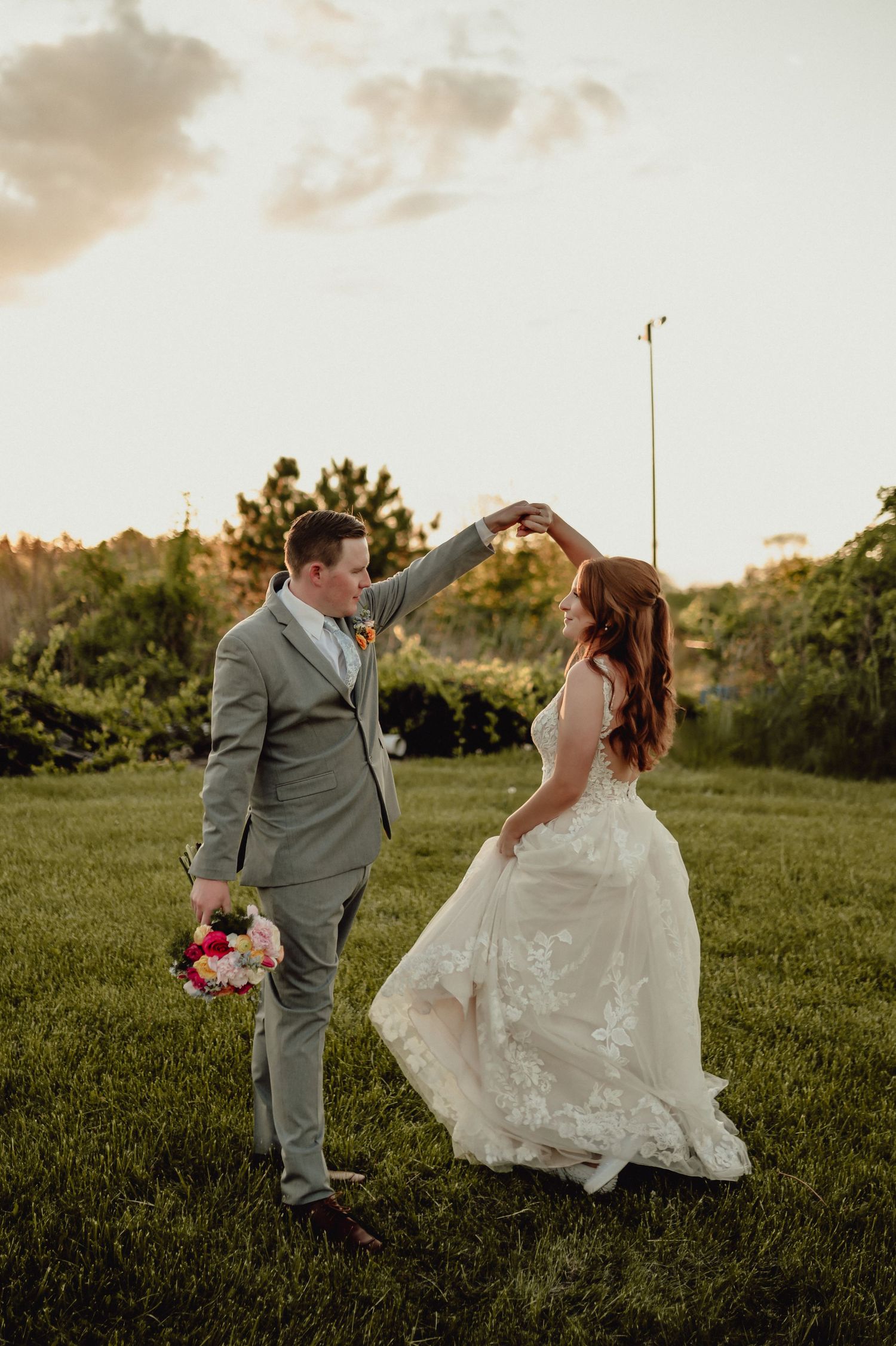 A couple in wedding attire dances playfully together in a grassy field at sunset.