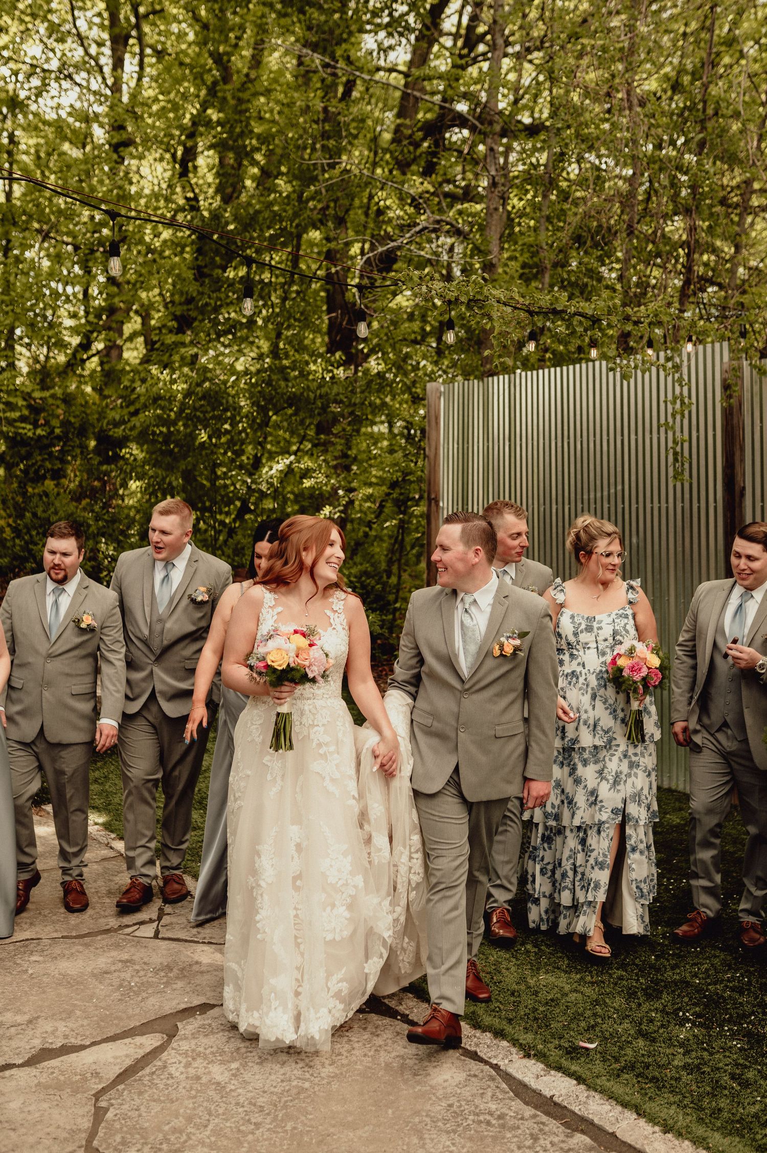 Wedding party in gray suits and floral dresses pose together outdoors in a garden setting.