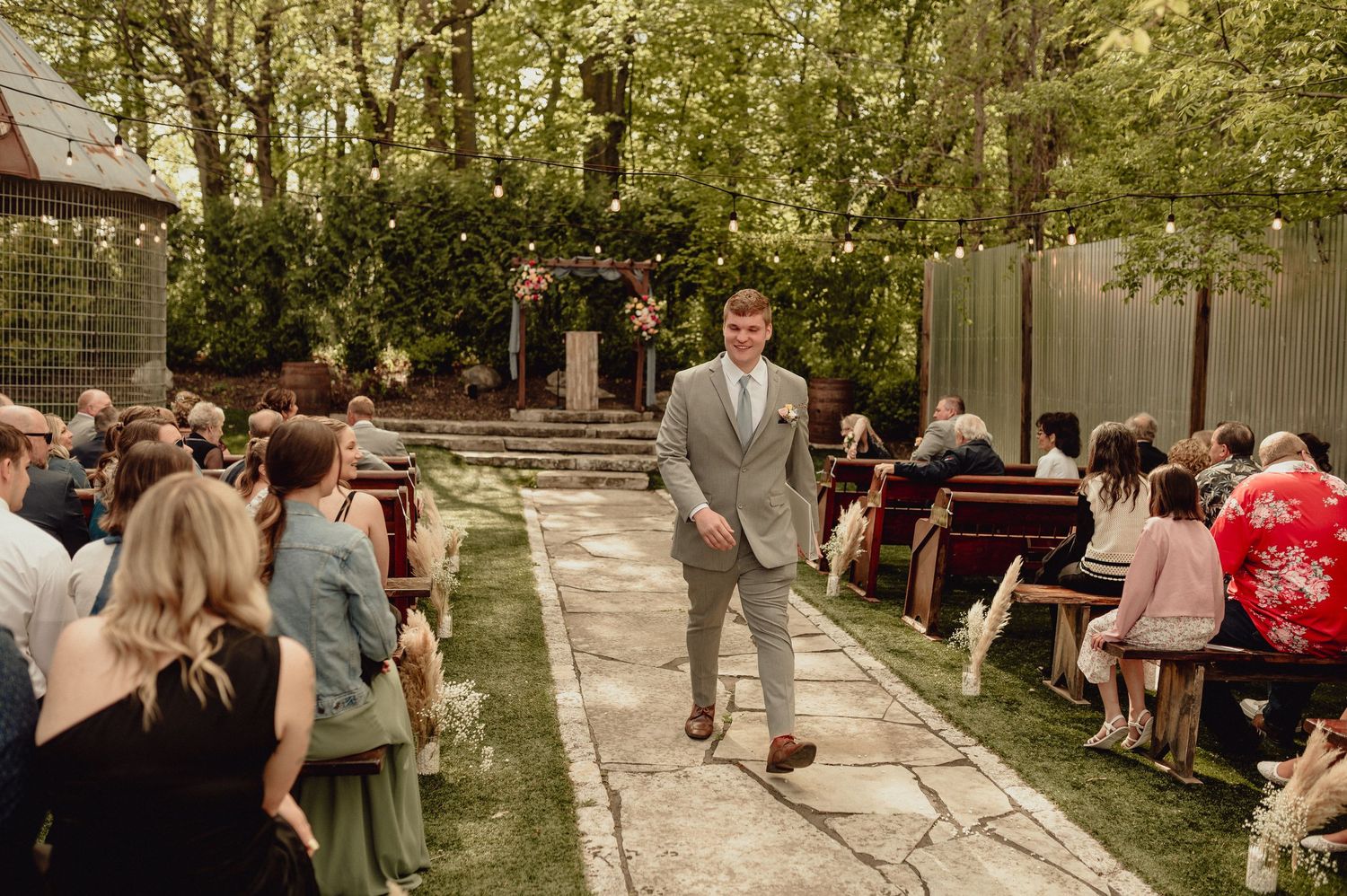 Wedding guests gather in an outdoor garden setting for a ceremony under tree canopies and string lights.