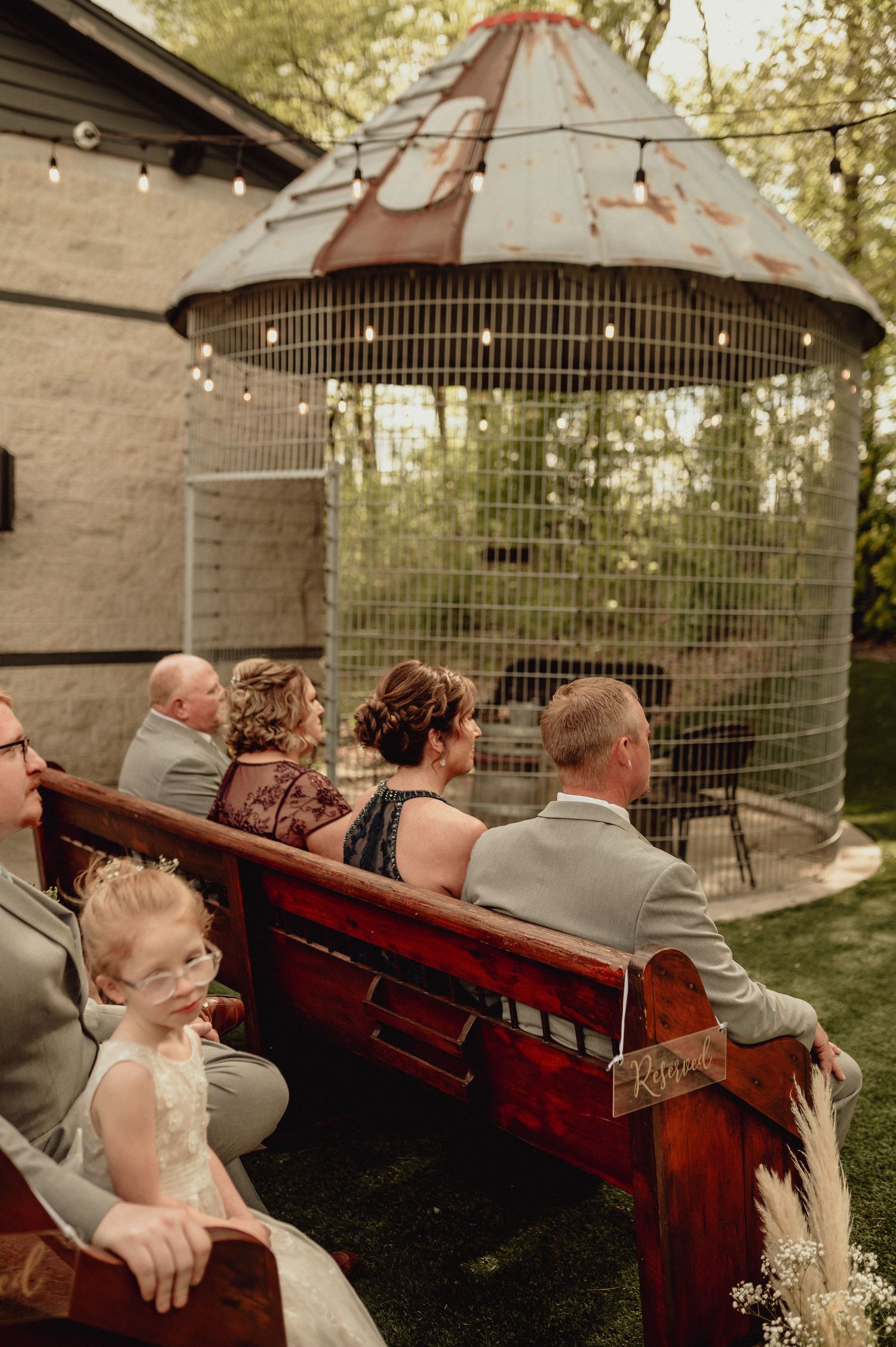 Wedding guests sit on wooden church pews outdoors beneath decorative metal gazebos during a garden ceremony.