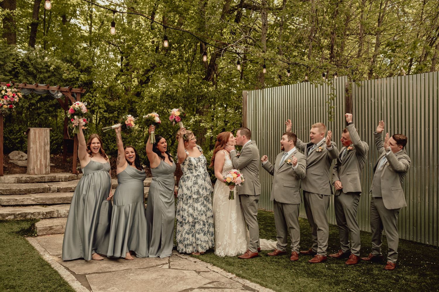 Wedding party in gray and floral dresses poses outdoors in a garden setting with natural greenery backdrop.