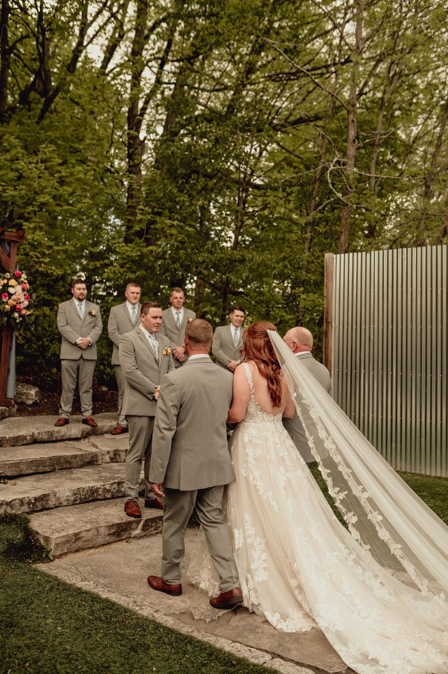 Wedding couple walks down stone pathway surrounded by wedding party in grey attire.