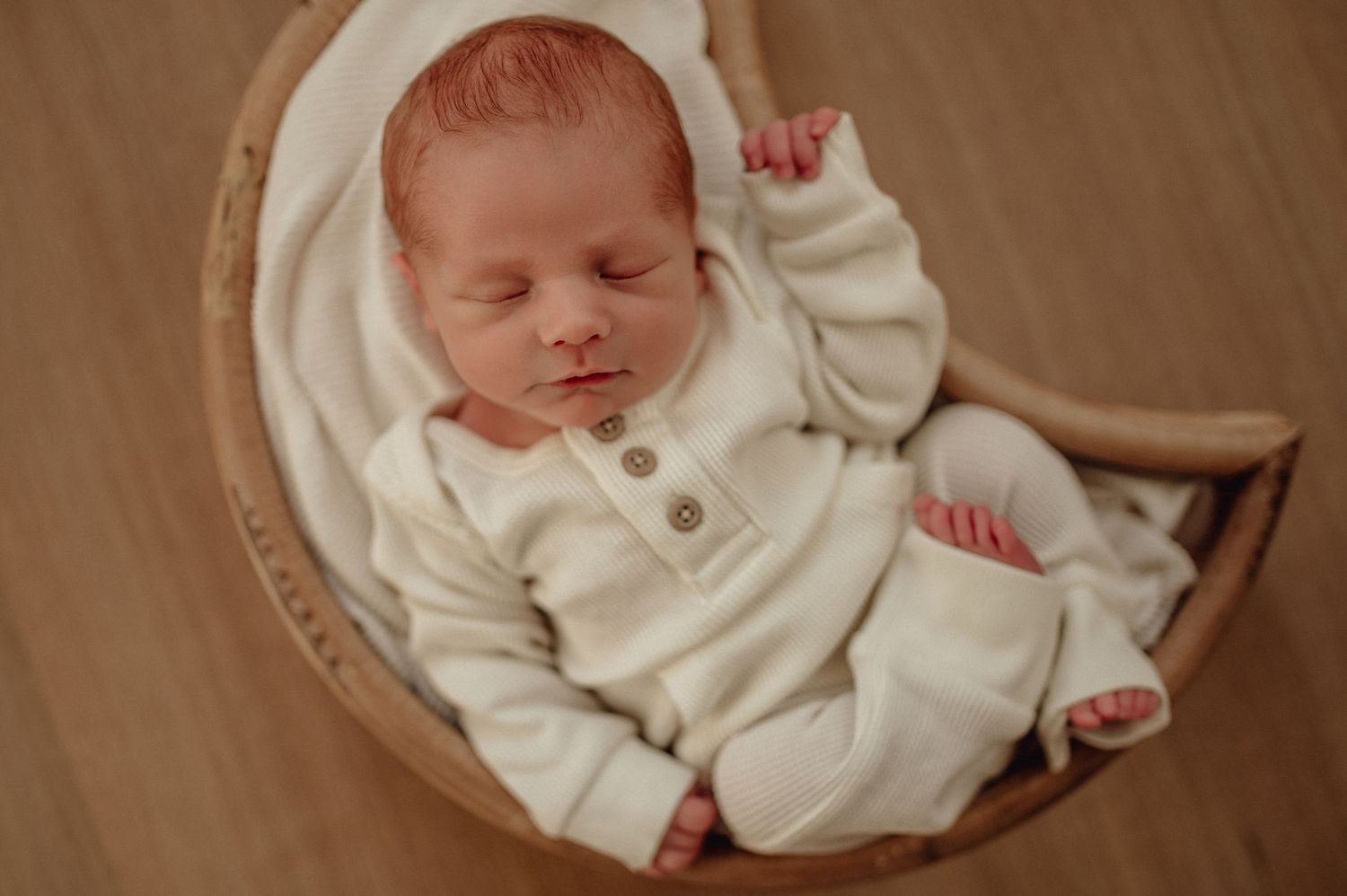 Sleeping newborn in cozy white outfit rests in wooden bowl.