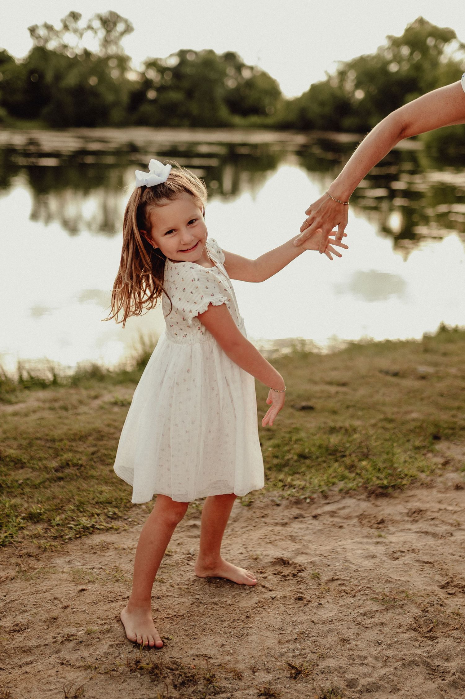 Young child in white dress twirling by a lakeside at sunset.