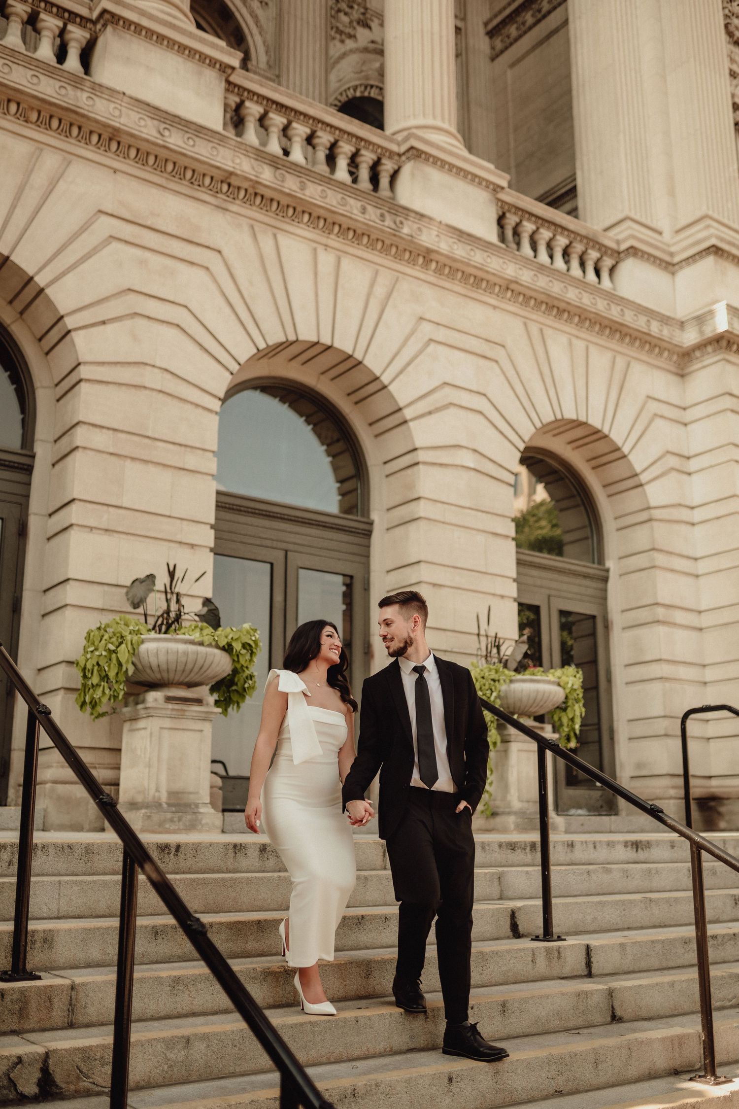 Wedding couple walking down grand stone steps of historic building with classical architecture.