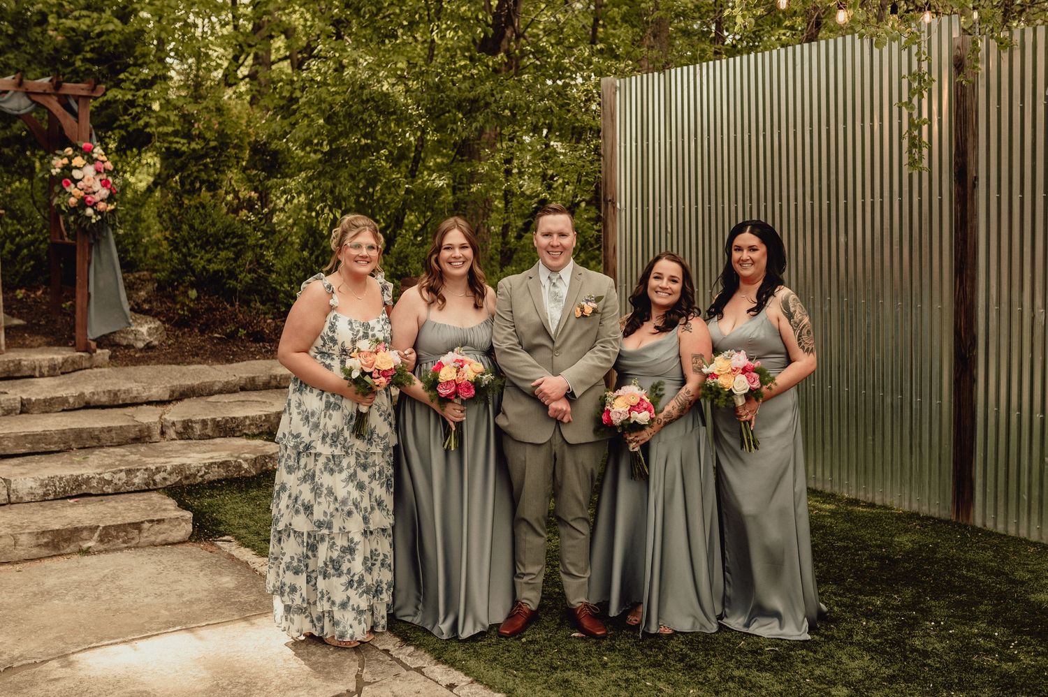 A group wearing sage green bridesmaid dresses poses together for outdoor wedding photos with coral bouquets.