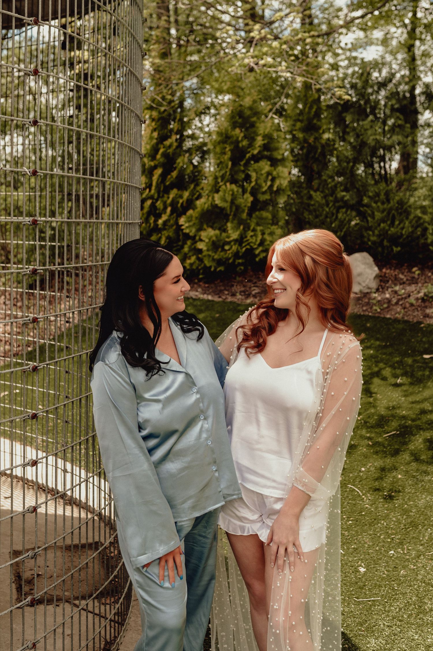 Two friends in contrasting outfits share a laugh together in a garden setting with metal fencing.