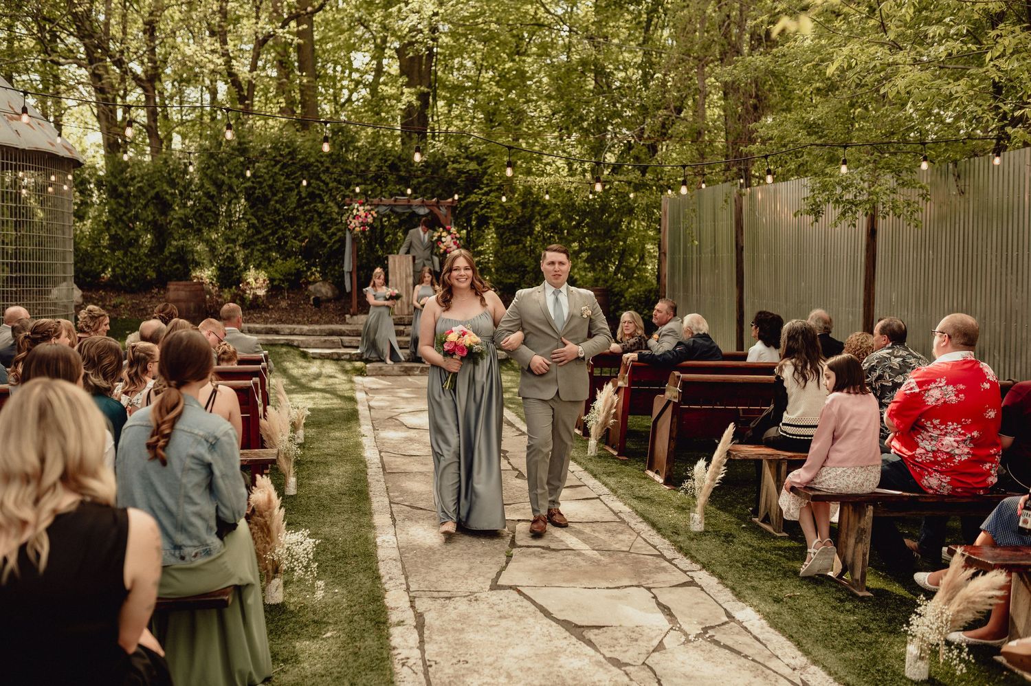 Wedding guests gather outdoors at picnic tables under trees during a casual backyard celebration.