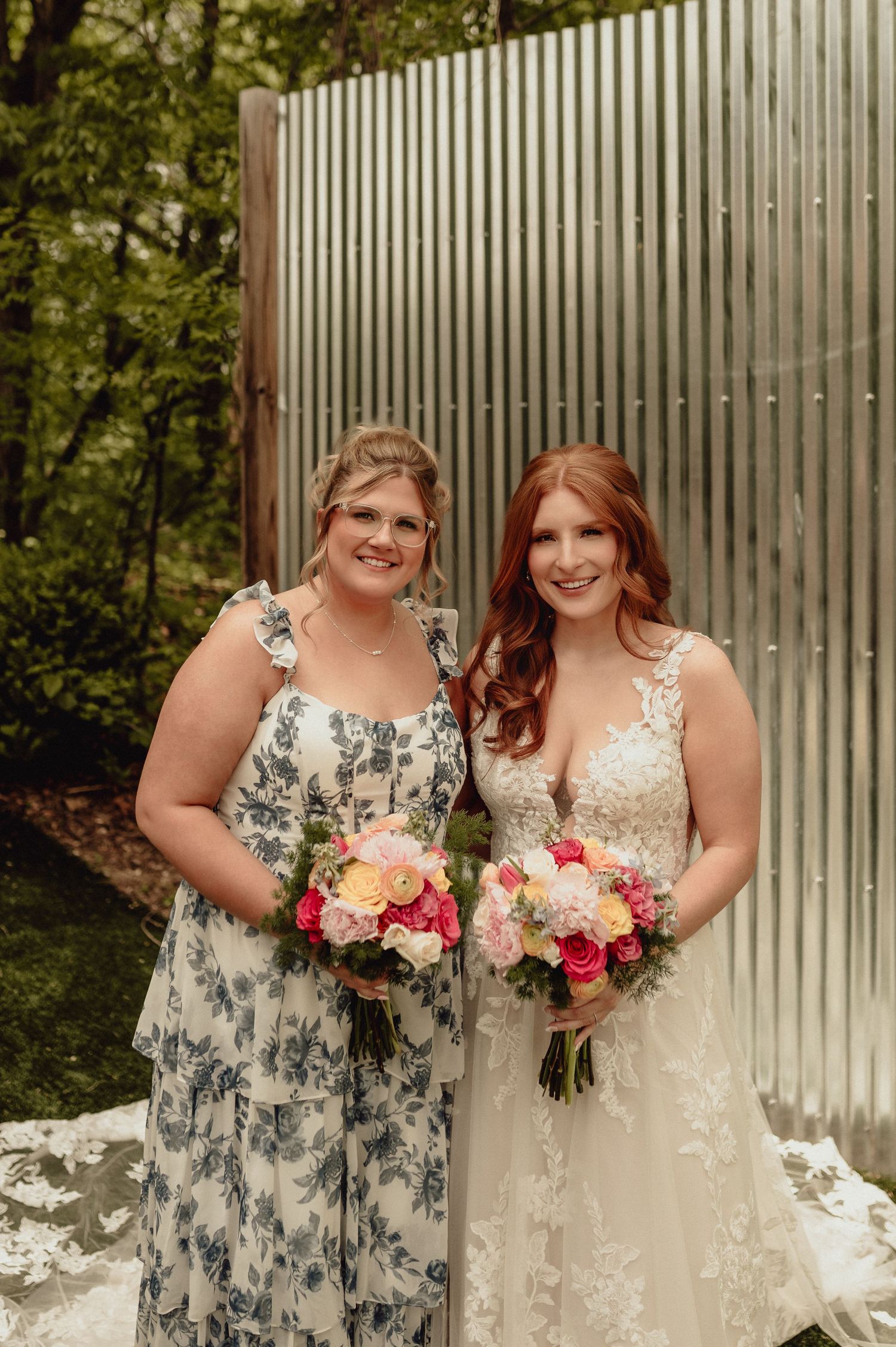 Two women in formal dresses pose together at an outdoor wedding venue with floral bouquets and a metal wall backdrop.