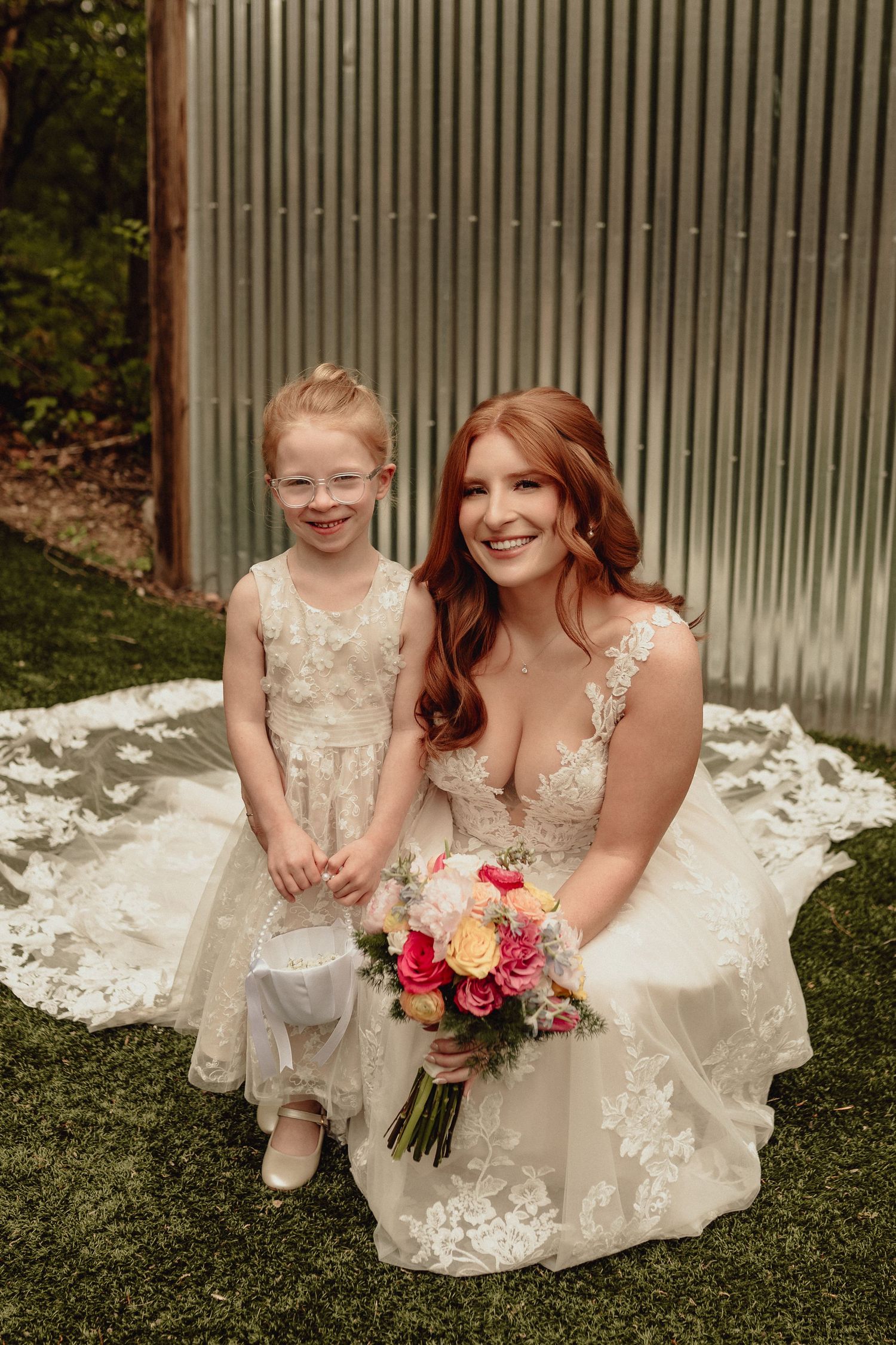 A bride in a lace wedding dress kneels on grass holding a pink and peach bouquet next to a young attendant in cream dress.
