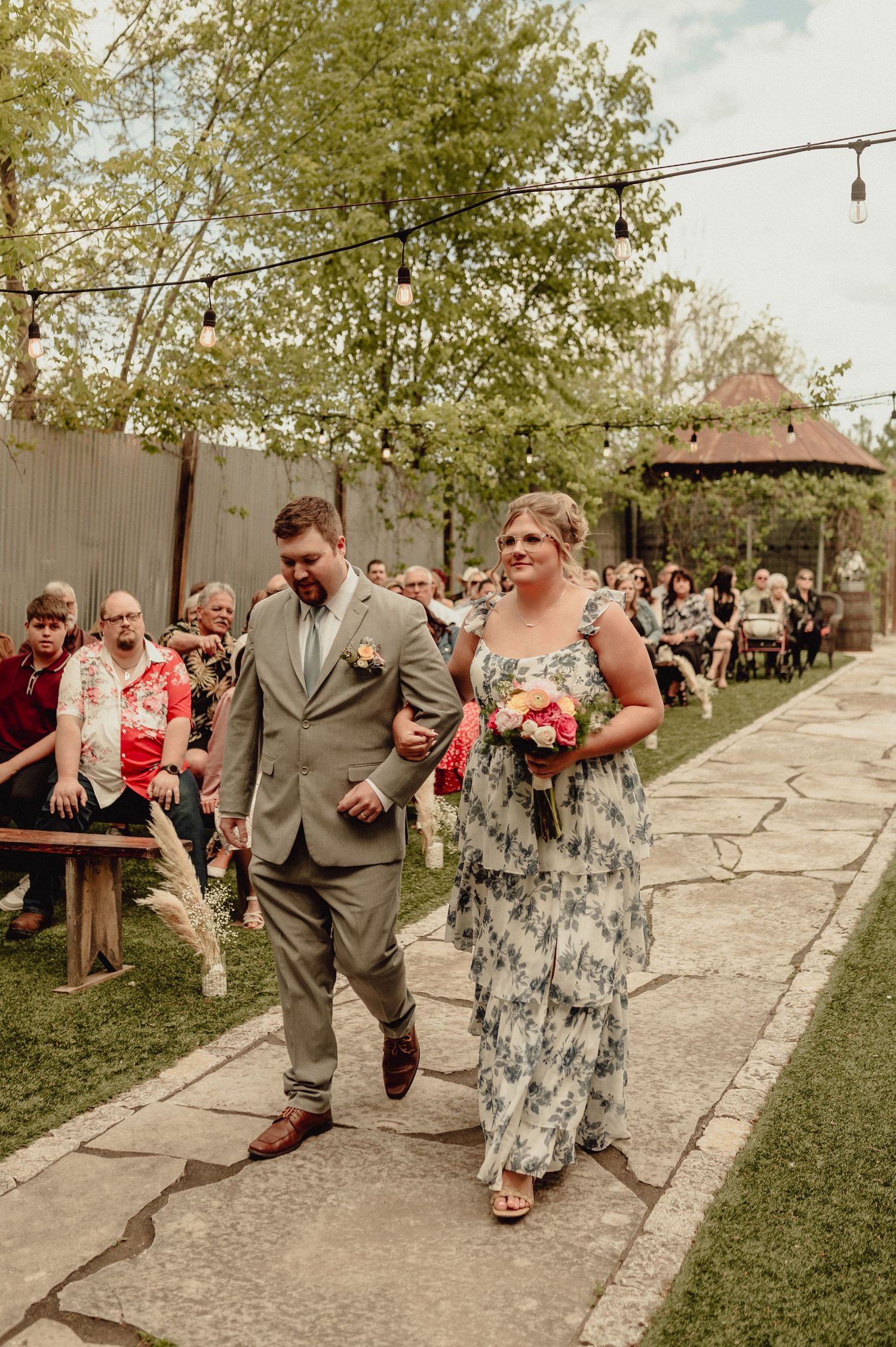 Wedding party members perform a fun synchronized dance move on a concrete path during an outdoor celebration.