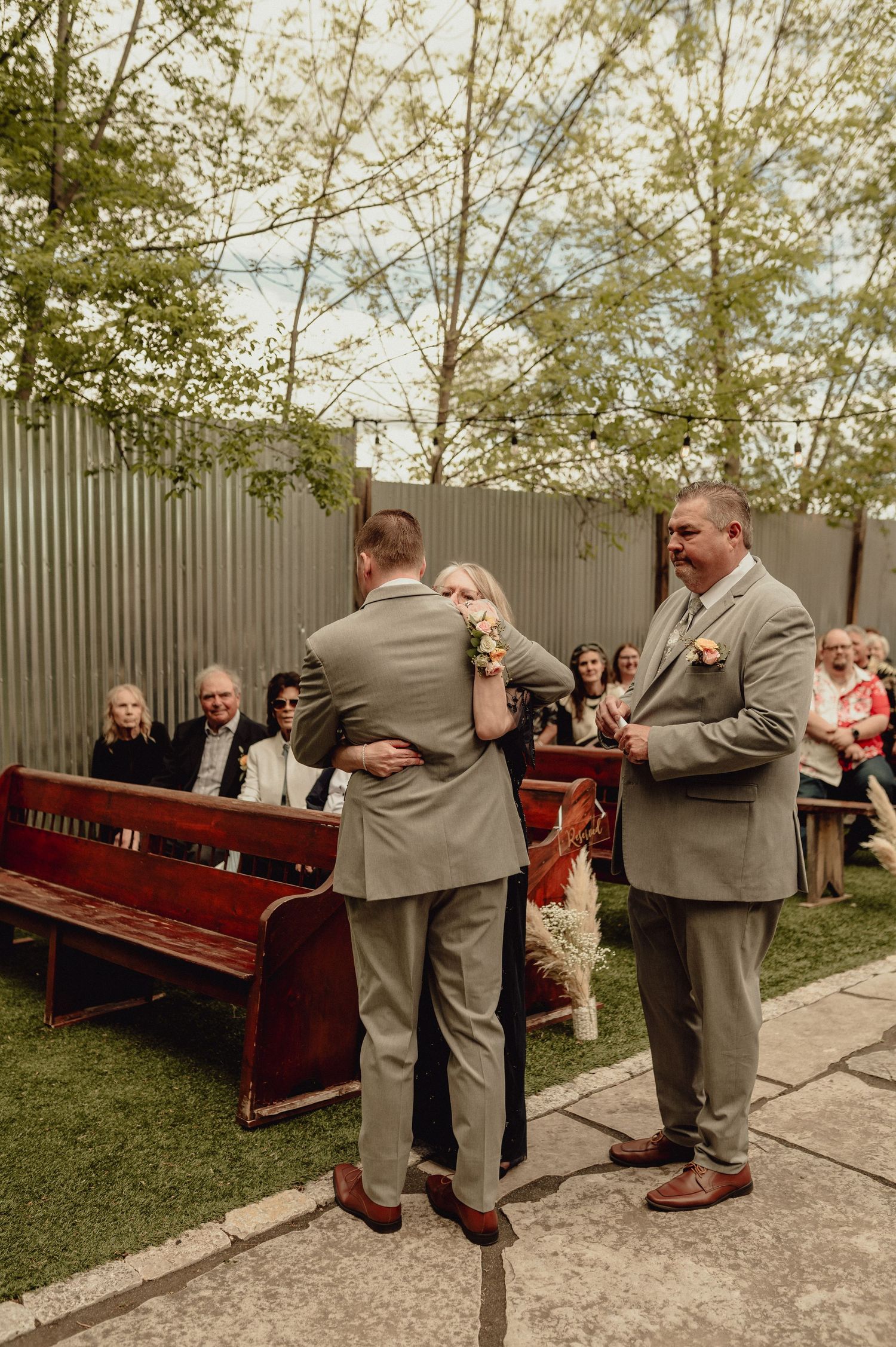 Outdoor wedding ceremony with wooden church pews and guests seated under trees behind a corrugated metal fence.