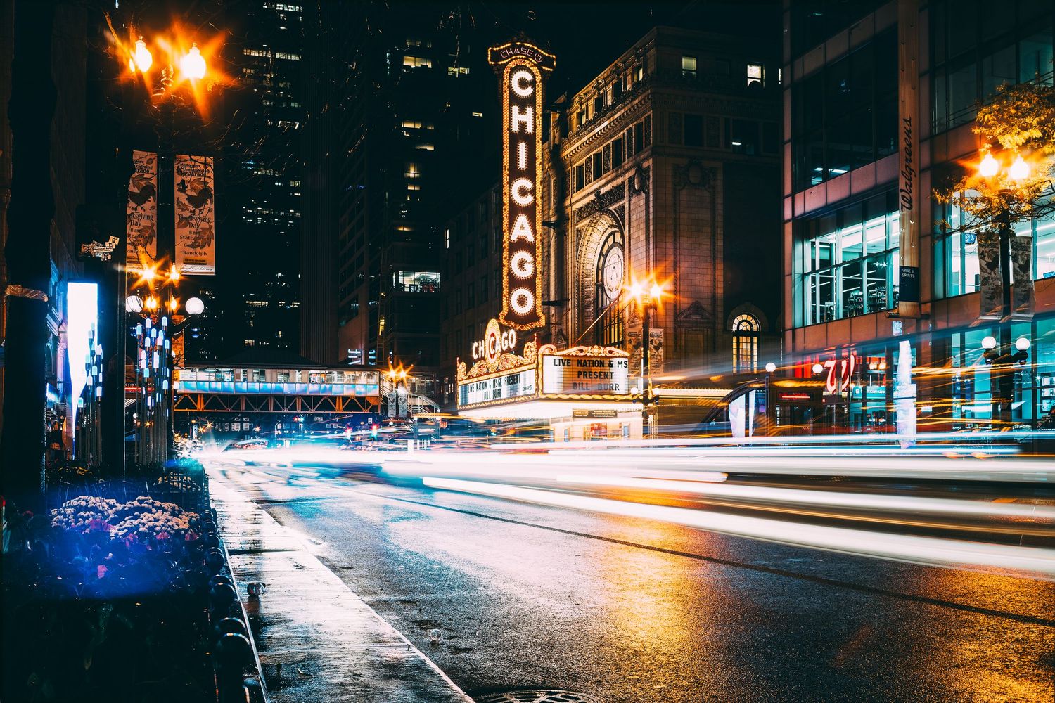 City street at night with light trails from passing vehicles and glowing street lamps.