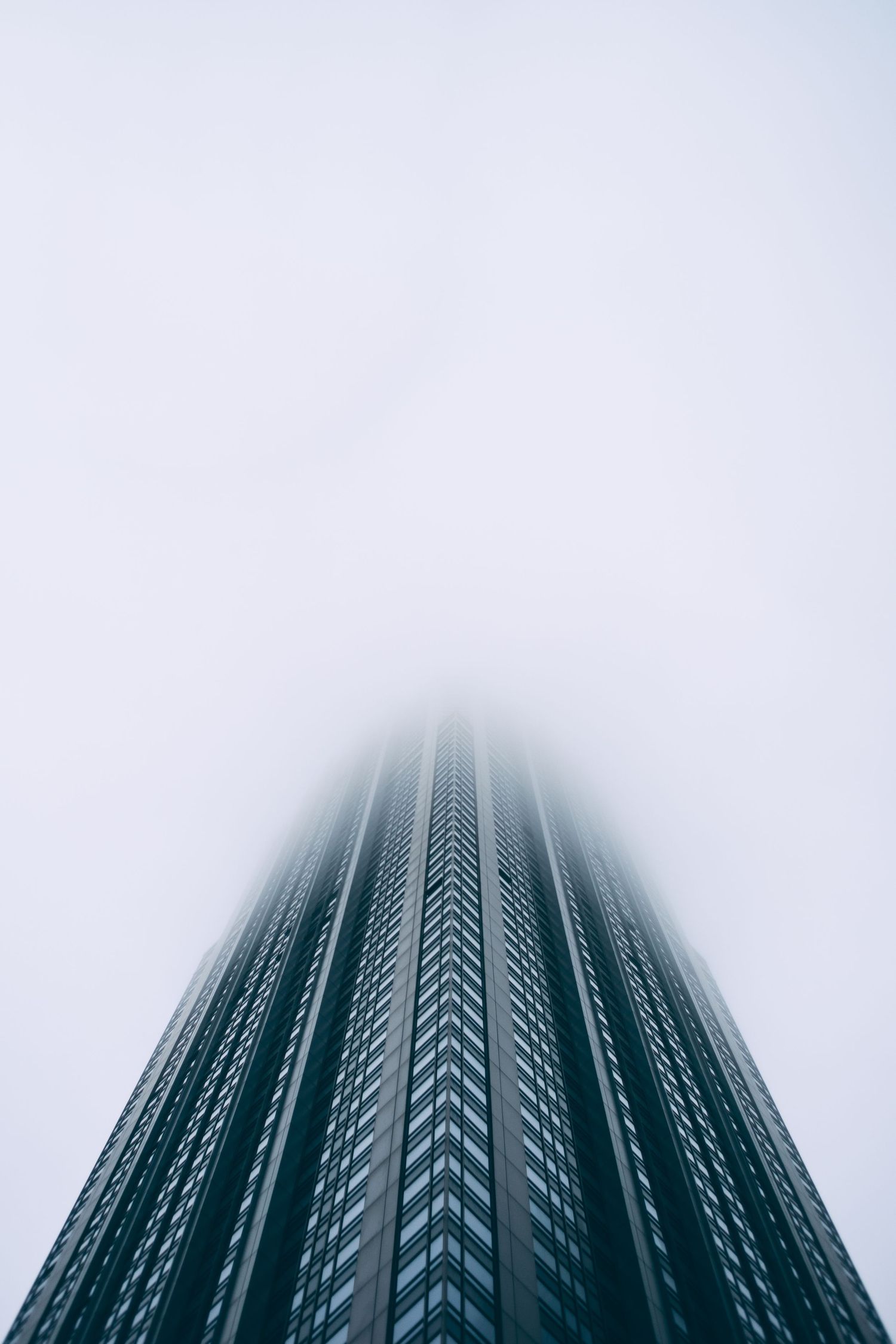 Modern skyscraper disappearing into fog viewed from below against white sky.