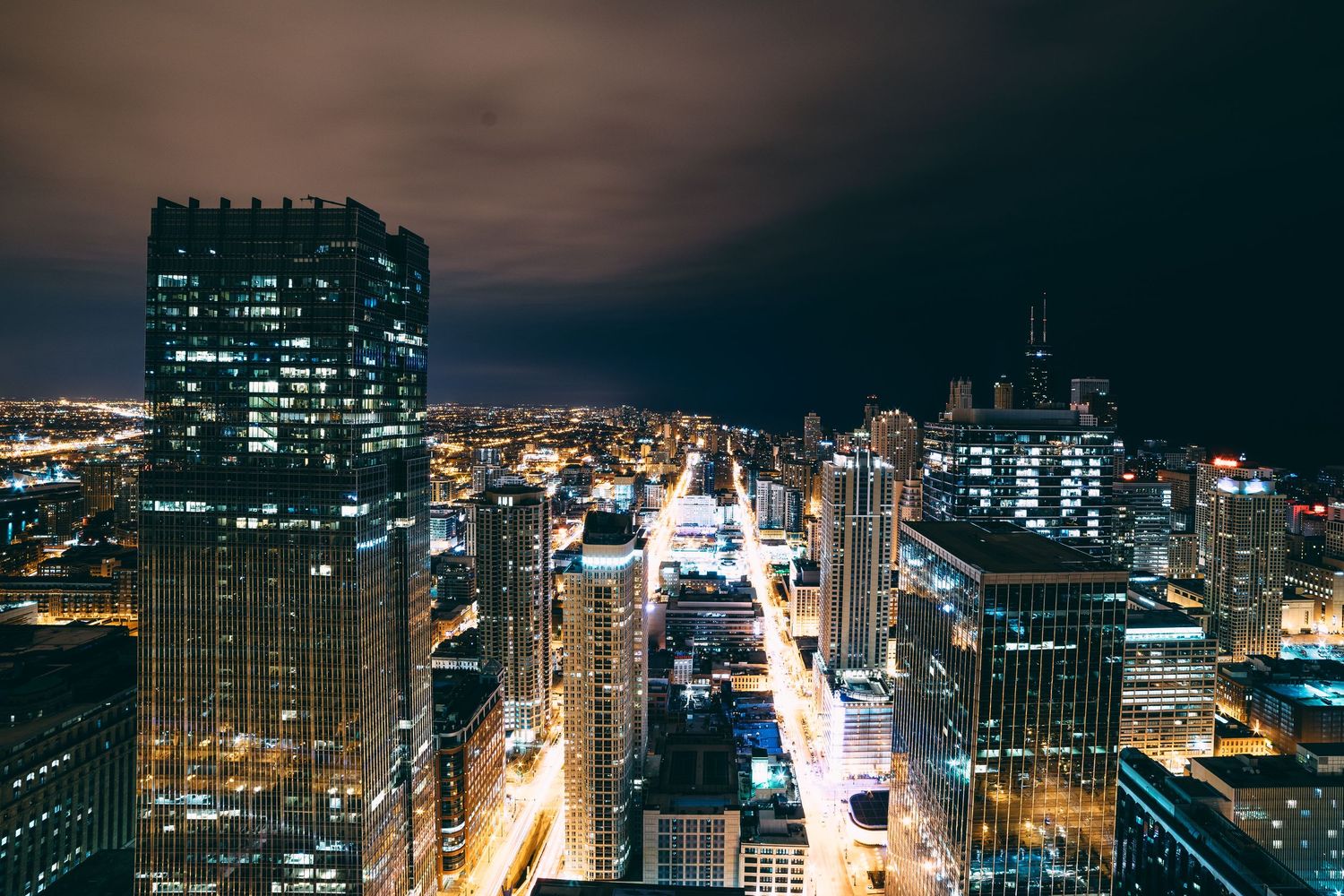 Nighttime cityscape showing illuminated skyscrapers and urban streets from above.
