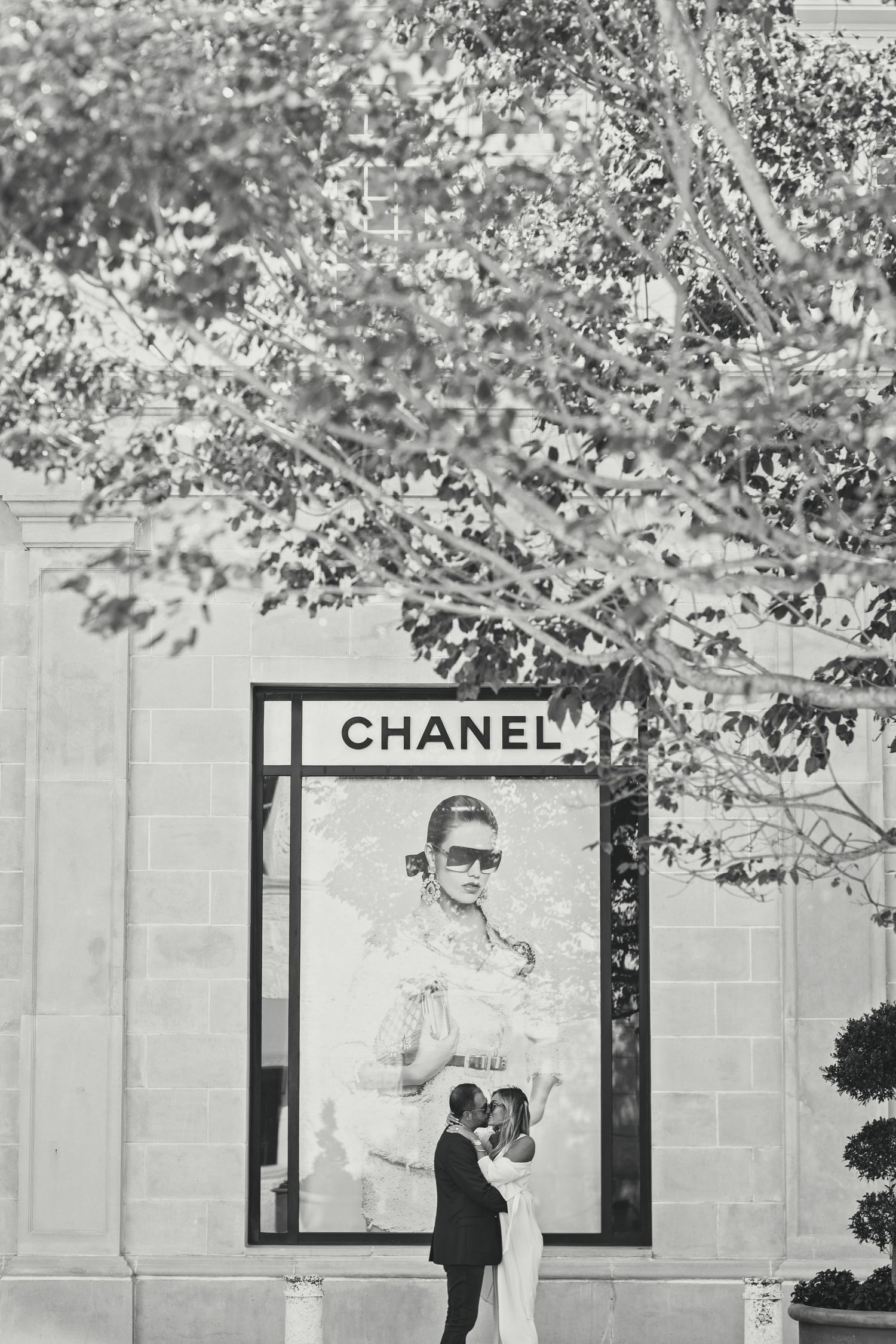 A couple shares an intimate moment outside a Chanel storefront with leafy branches overhead.