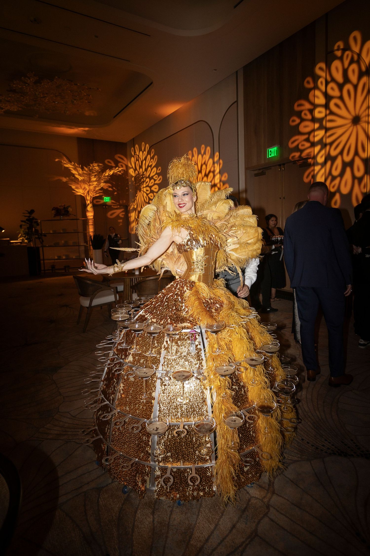 Performer in an elaborate gold ball gown dress serves champagne at an elegant event with decorative lighting.