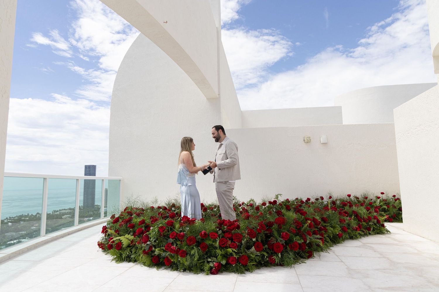 Couple holding hands on a rooftop terrace surrounded by red roses with ocean views and white architectural arches.