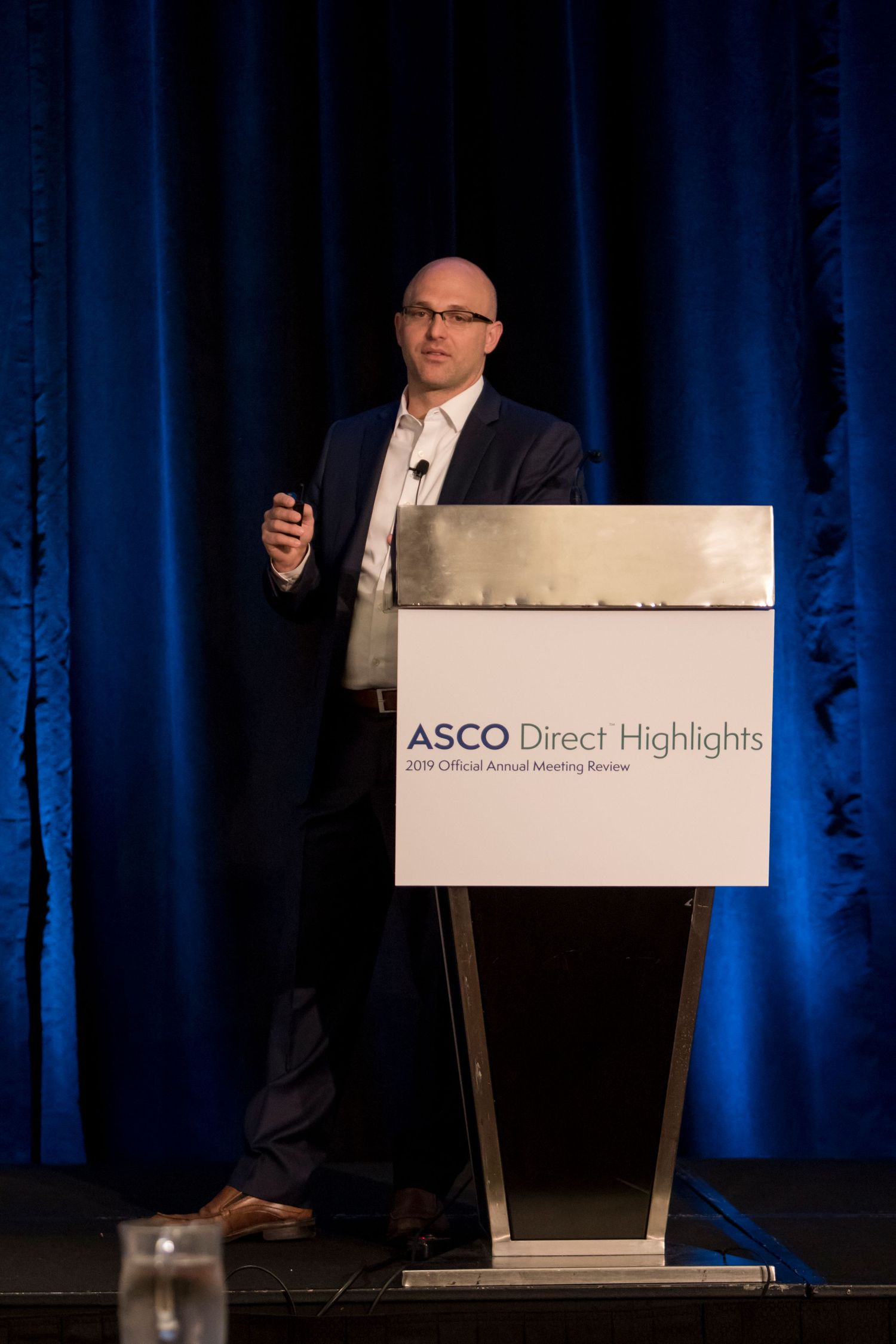 A speaker presenting at a podium with ASCO Direct Highlights text during a medical conference with blue curtains.