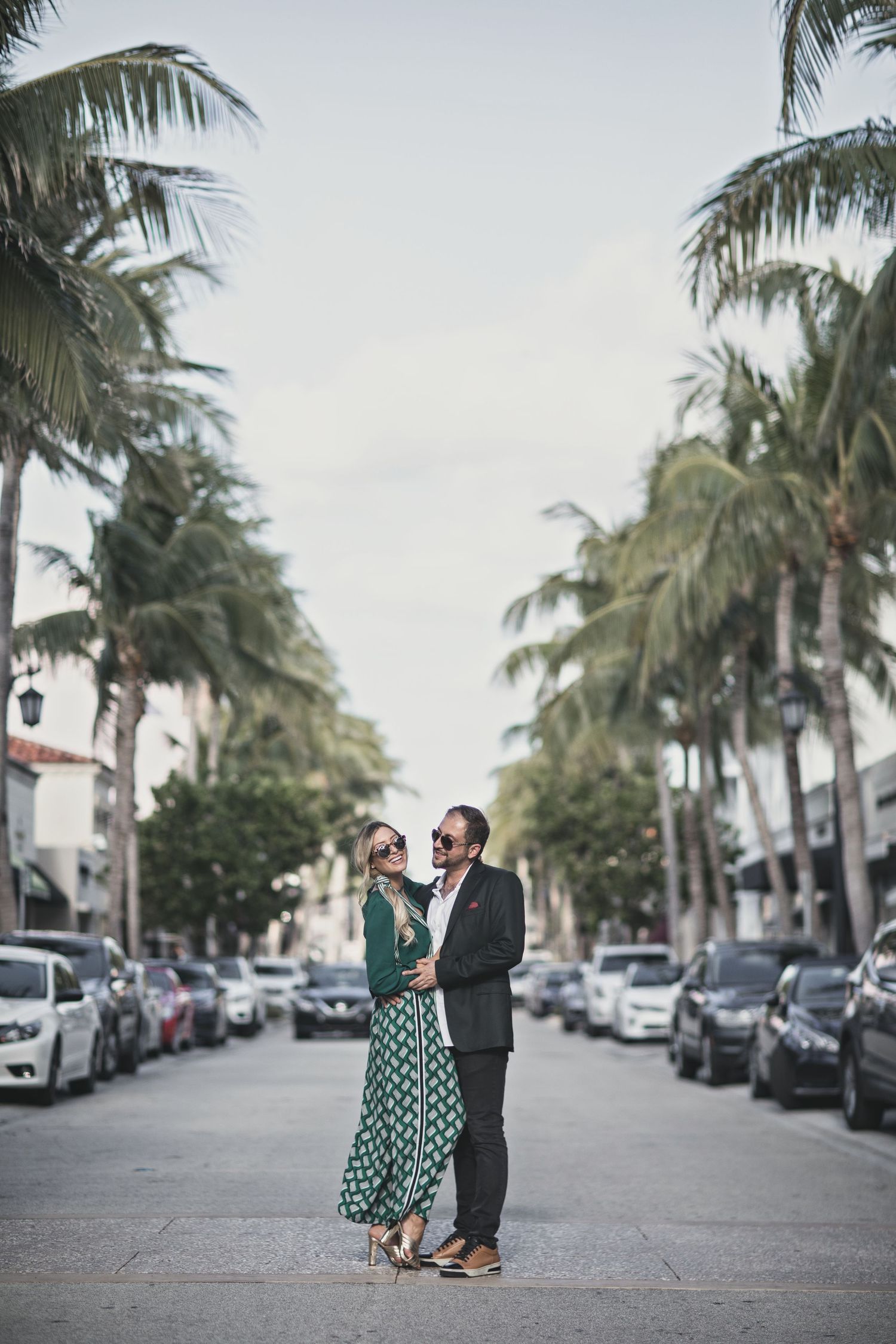 A couple stands on a palm tree-lined street wearing formal attire on a cloudy day.