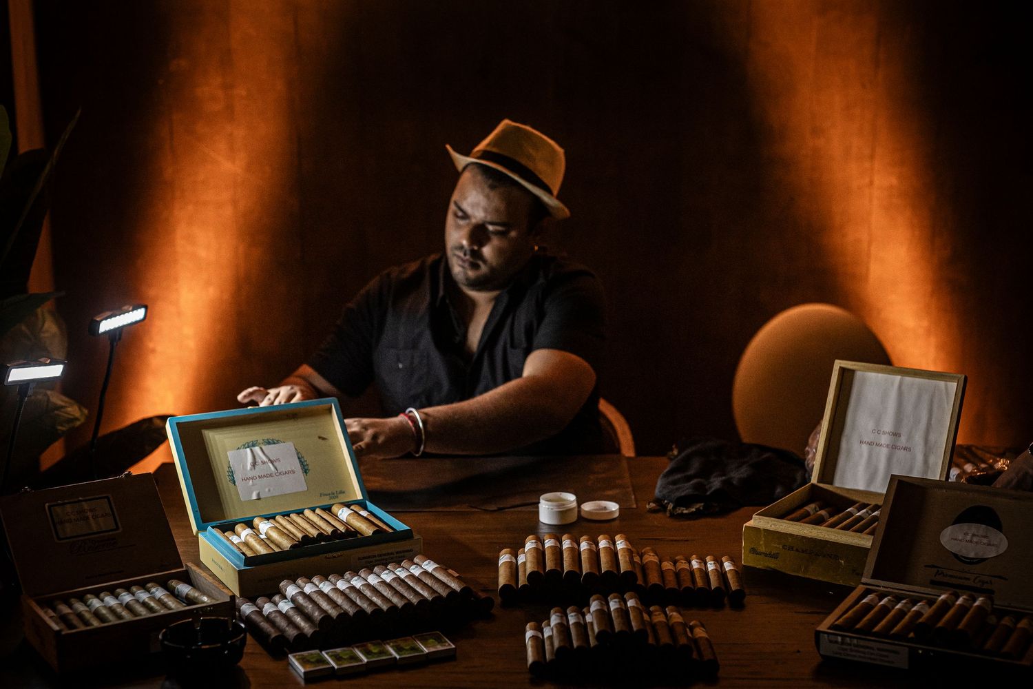 A person in a black shirt and brown hat sits at a table with neatly arranged cigar boxes and accessories in moody lighting.