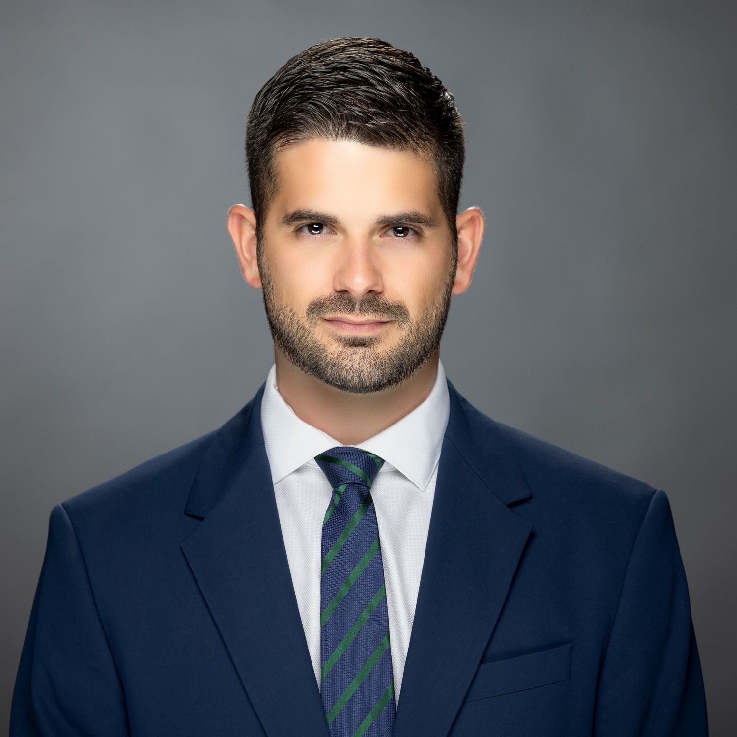 Professional headshot of a suited person wearing a navy blazer and striped tie against a gray background.