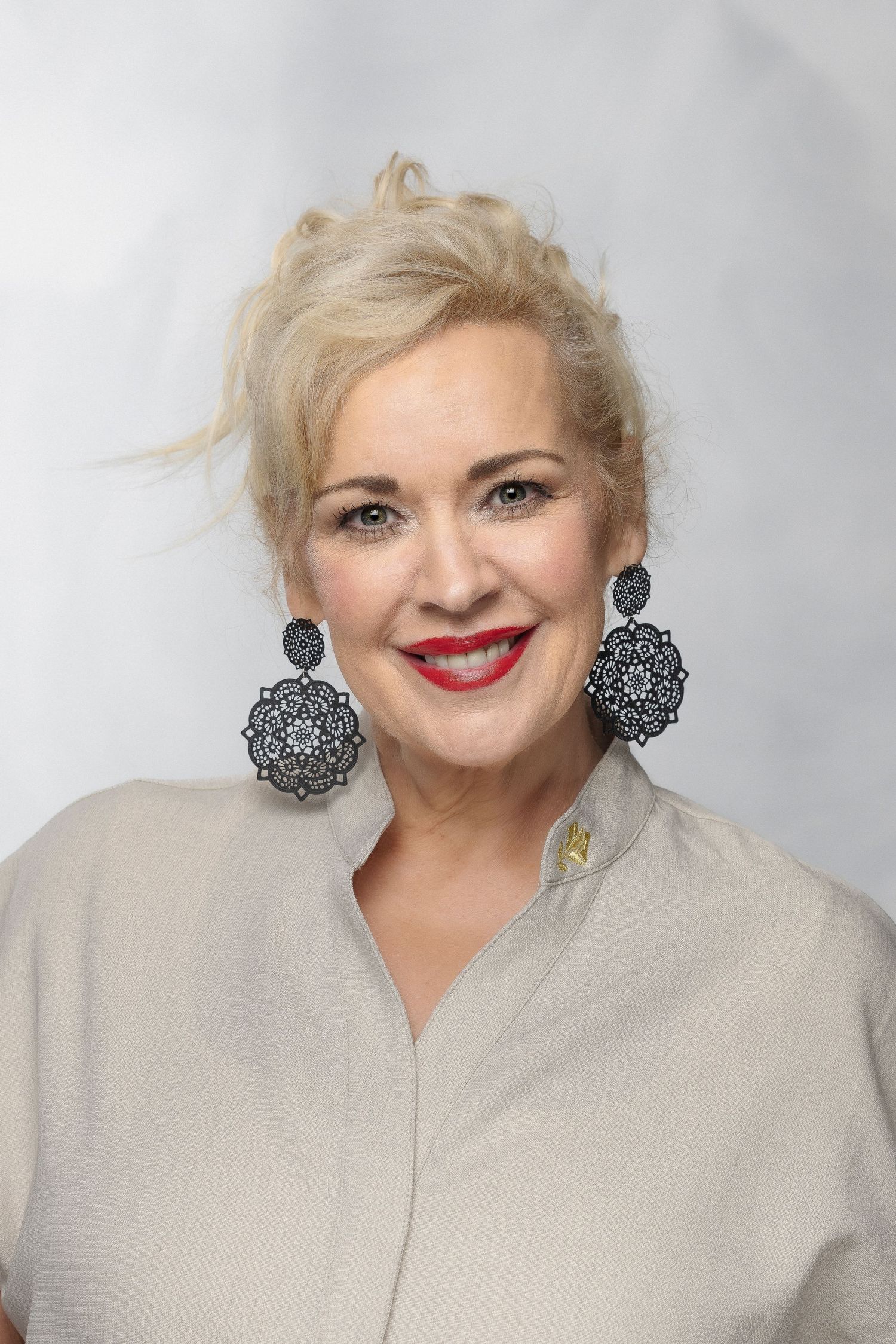 A person wearing a light grey top and black lace earrings smiles warmly while posing against a grey background.