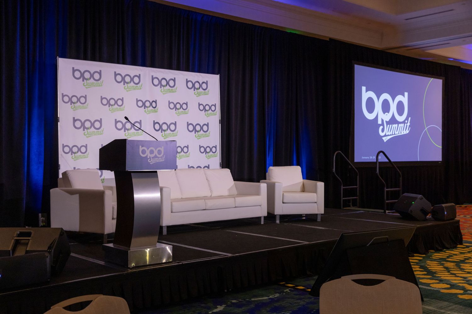 Professional conference stage setup with white furniture and blue lighting on wide screen displays.