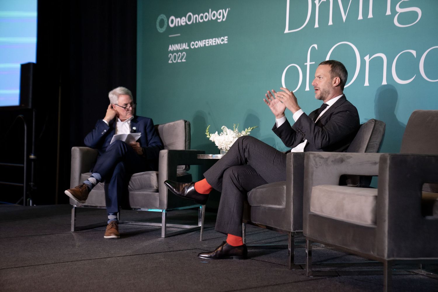 Conference stage setup features two armchairs and a small table against a teal-colored backdrop with lighting.