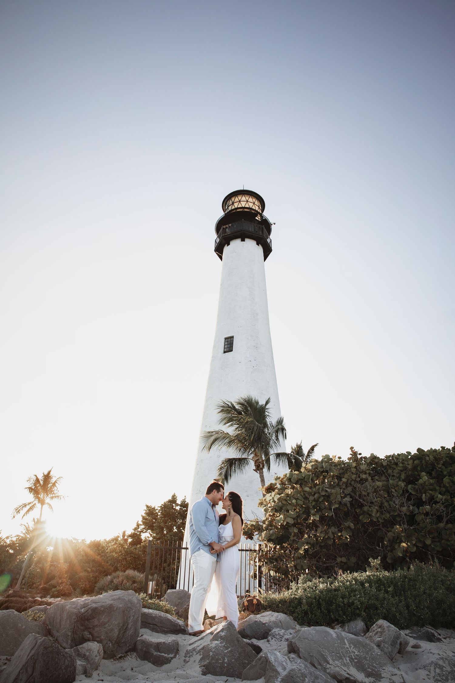 Classic white lighthouse stands tall against a bright sky with palm trees and rocks below.