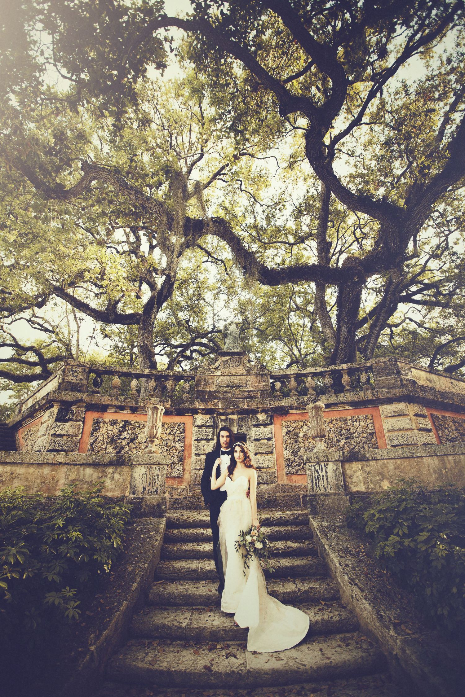 Wedding photo captured against rustic brick wall under overhanging trees.