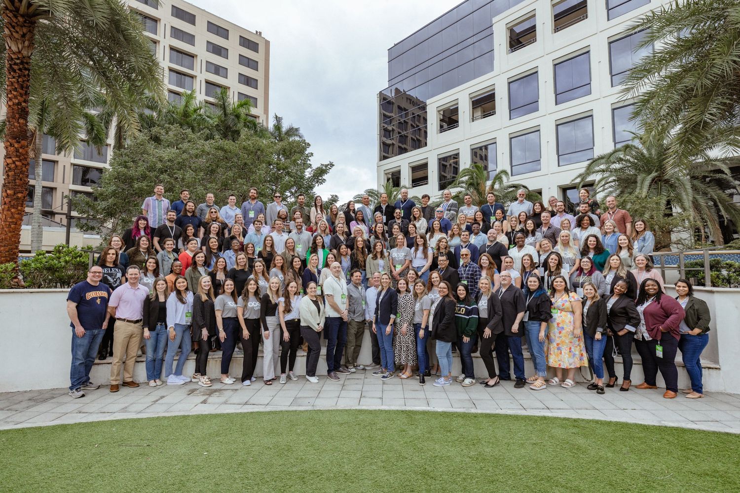 Large group of people gathered for a casual outdoor photo session in front of modern office buildings with palm trees.