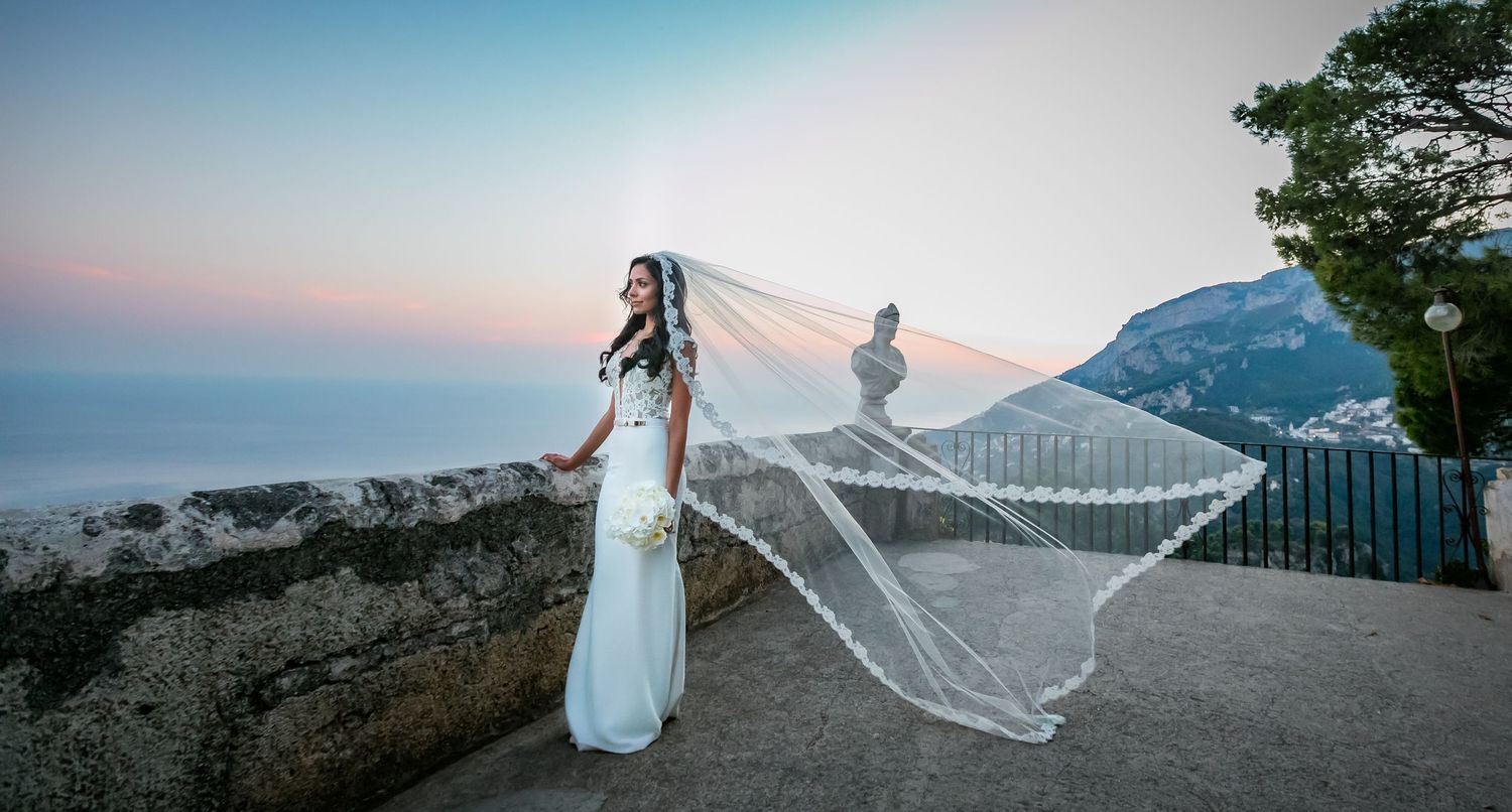 Long flowing veil catches wind against stunning mountain coastline view at sunset during wedding photoshoot.