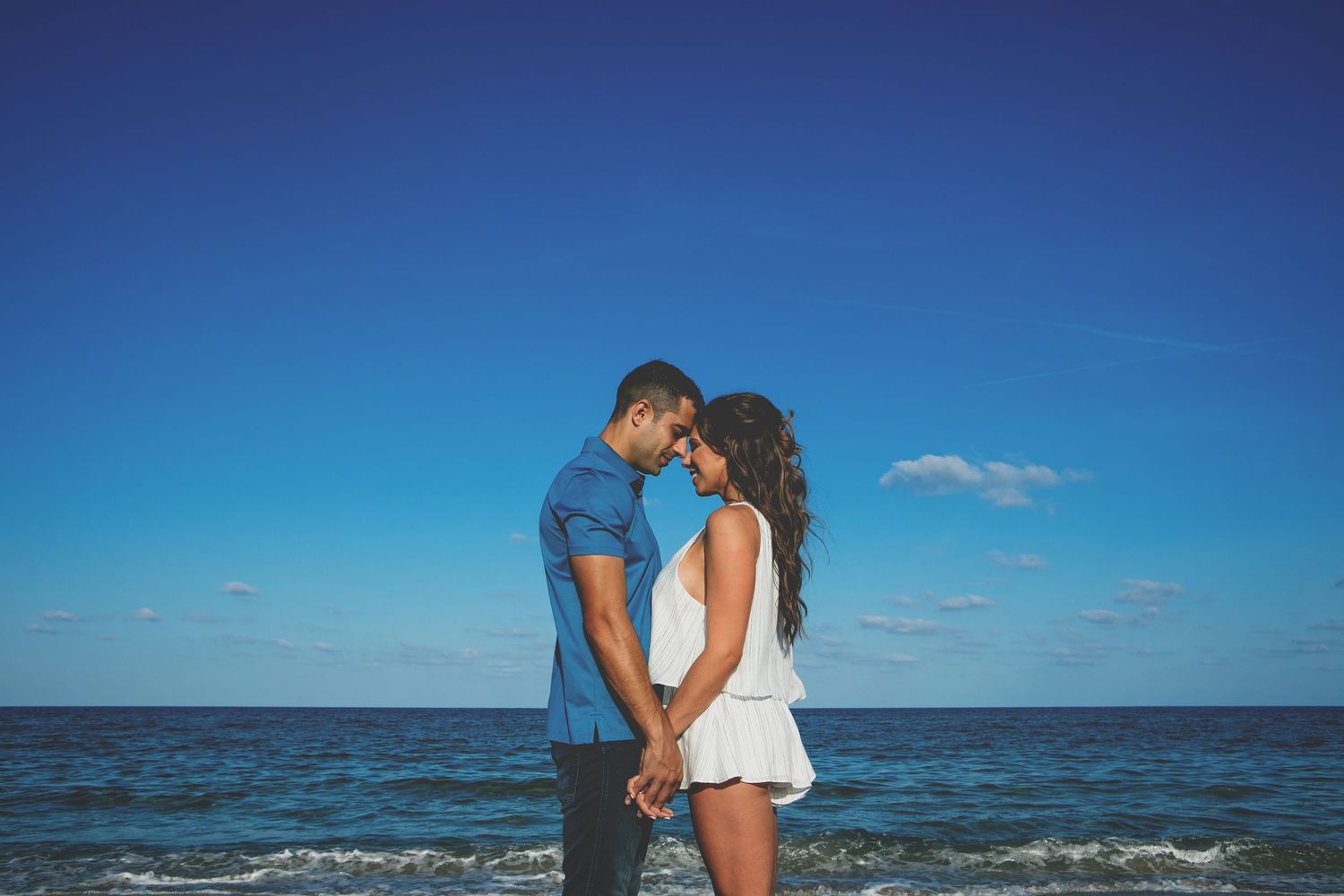 Romantic couple silhouetted against blue ocean horizon on tropical beach at sunset.