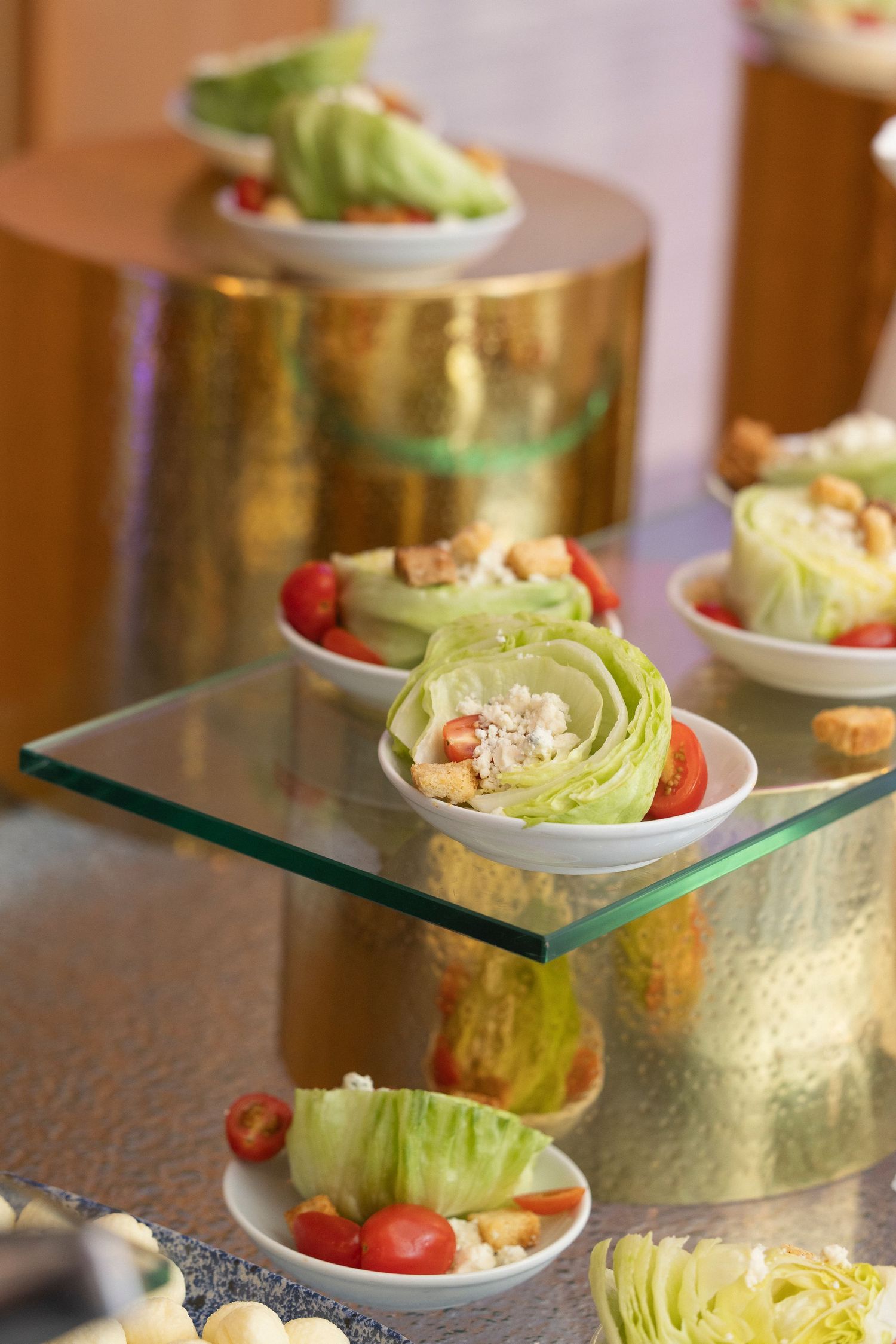 Catered food display featuring rows of iceberg lettuce wedge salads garnished with tomatoes on gold stands.