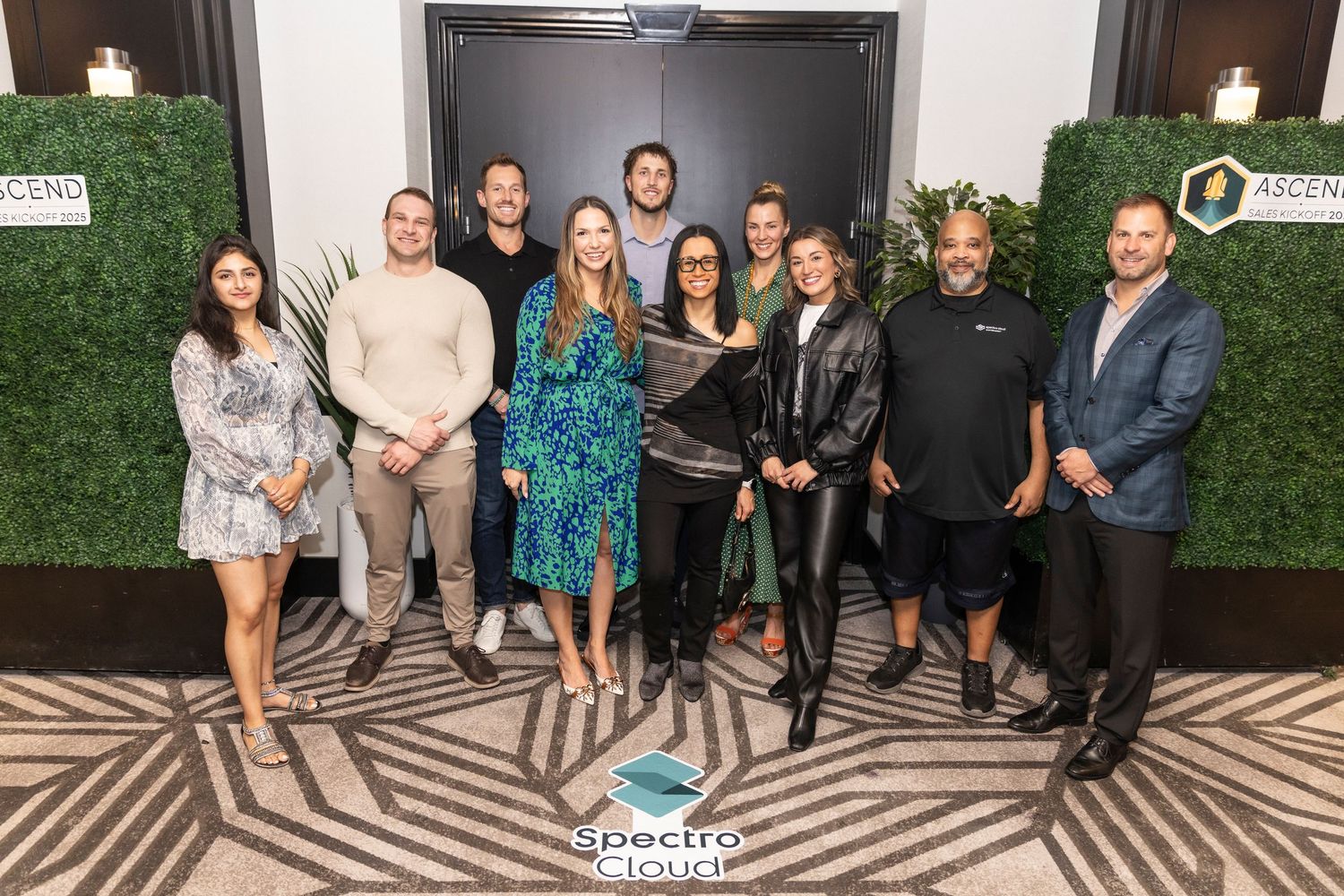 Group photo of alumni and guests posing together against a wall with green foliage decor at an event.