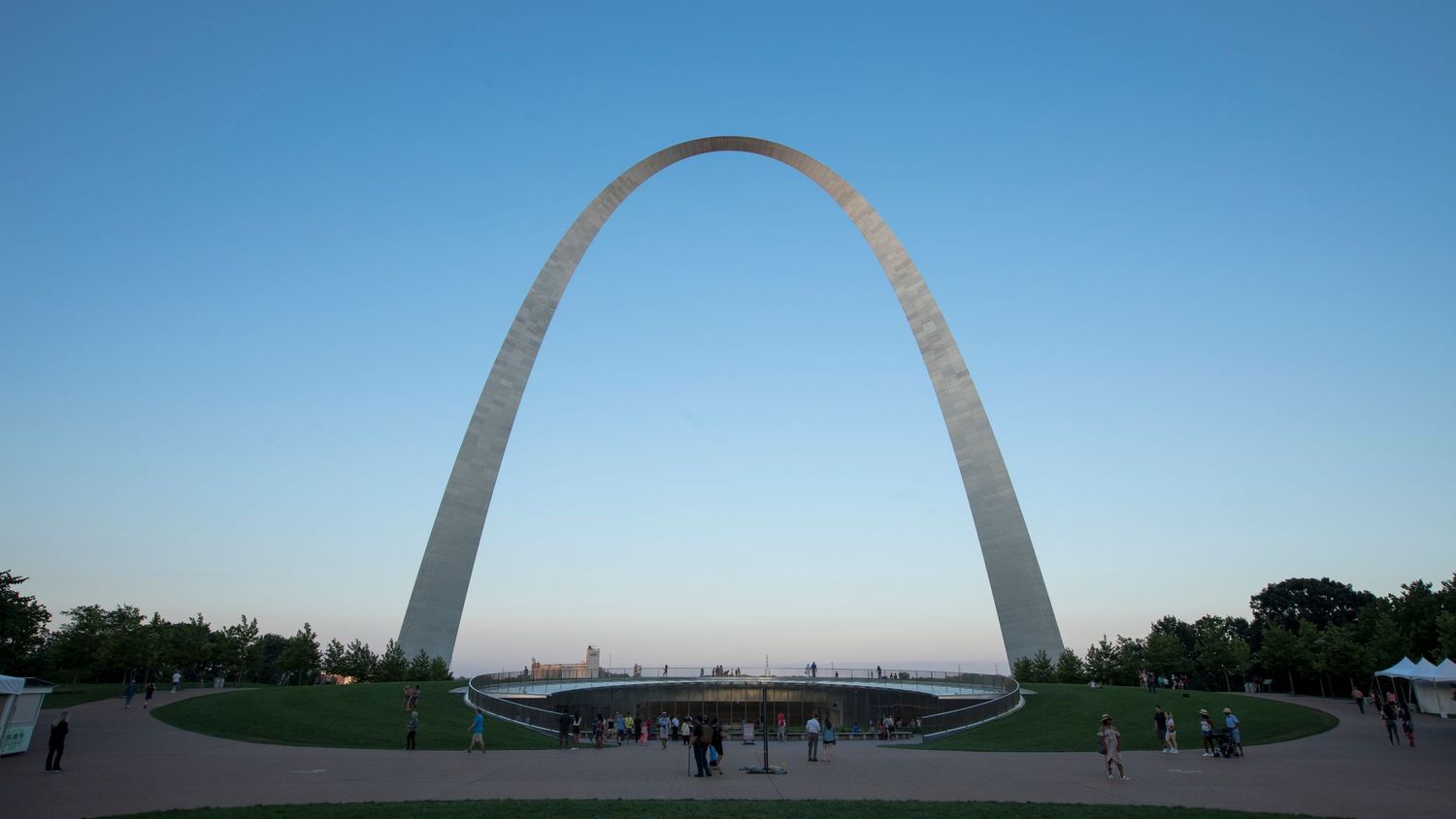 Panoramic view of the iconic Gateway Arch in St. Louis against a blue sky with scattered visitors on the surrounding grounds.