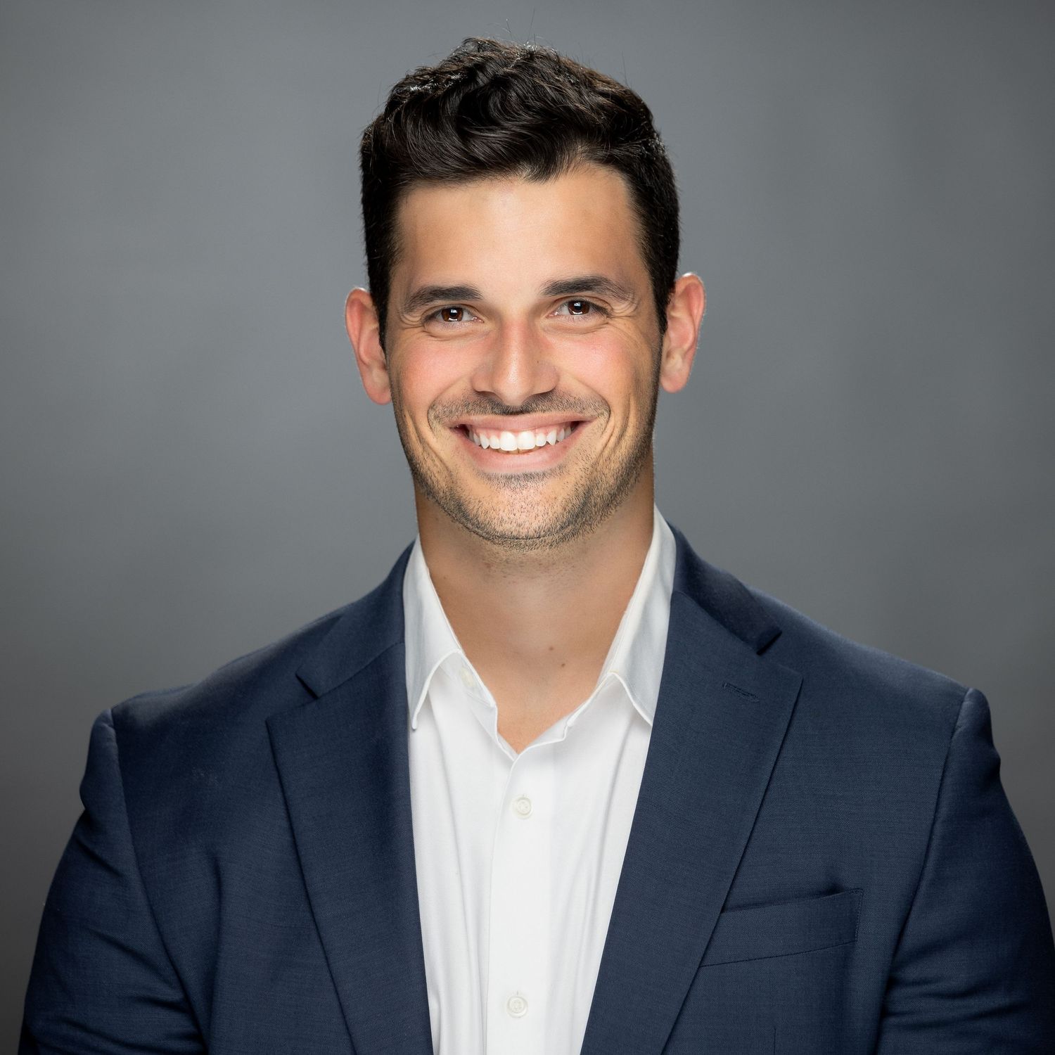 Professional headshot of someone wearing a navy suit jacket and white dress shirt with a bright confident smile.