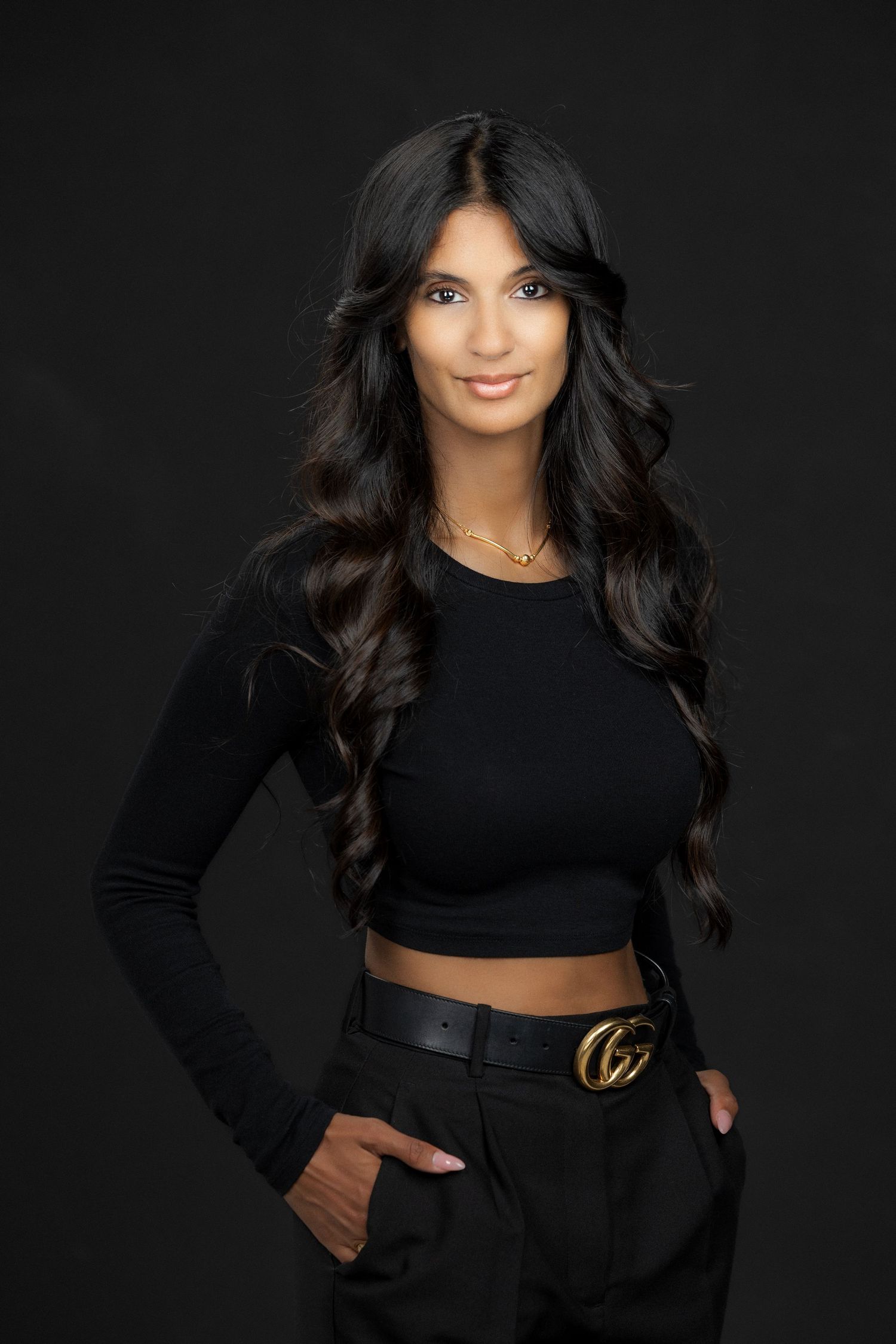 Fashion model wearing black crop top and leather belt with designer logo against dark background.