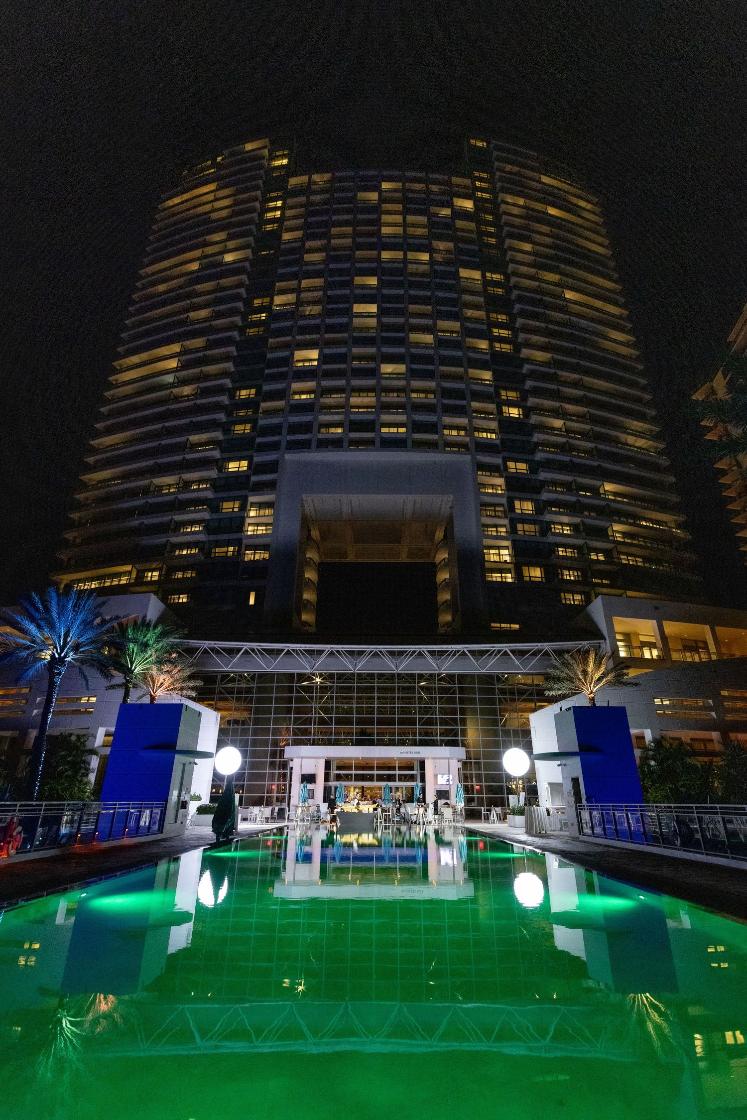 Night view of illuminated hotel building with green-lit outdoor deck and glowing palm trees at dusk.