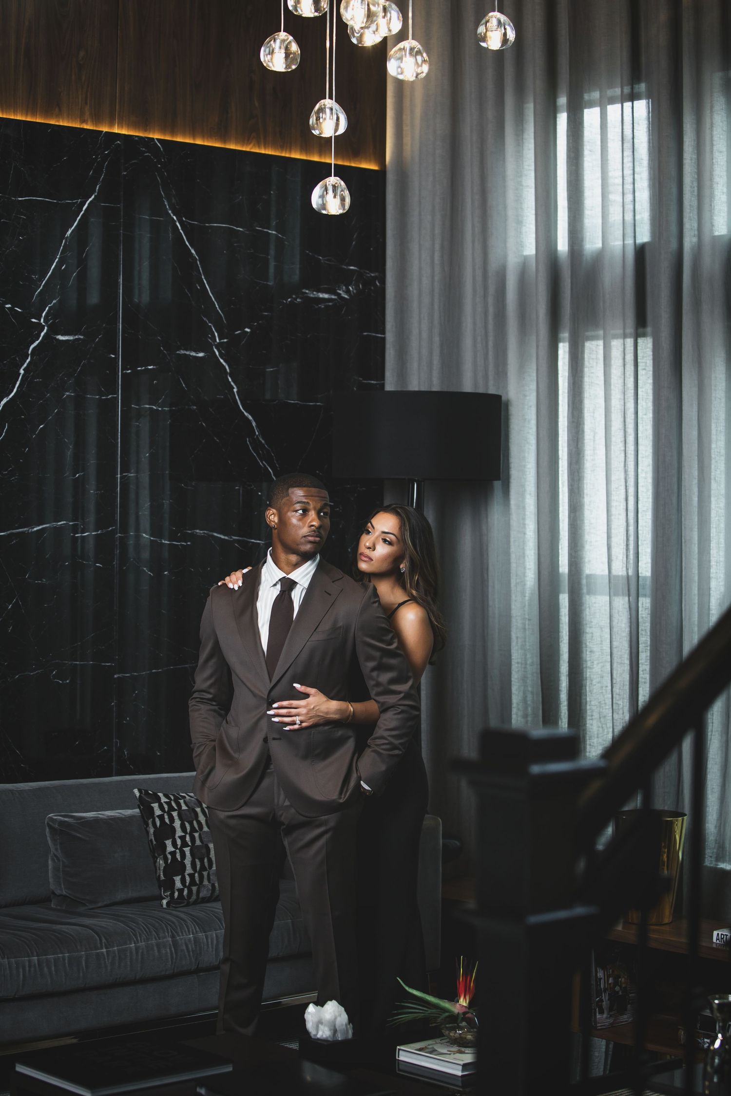 Elegant couple posing in modern room with black marble walls and hanging glass lights.