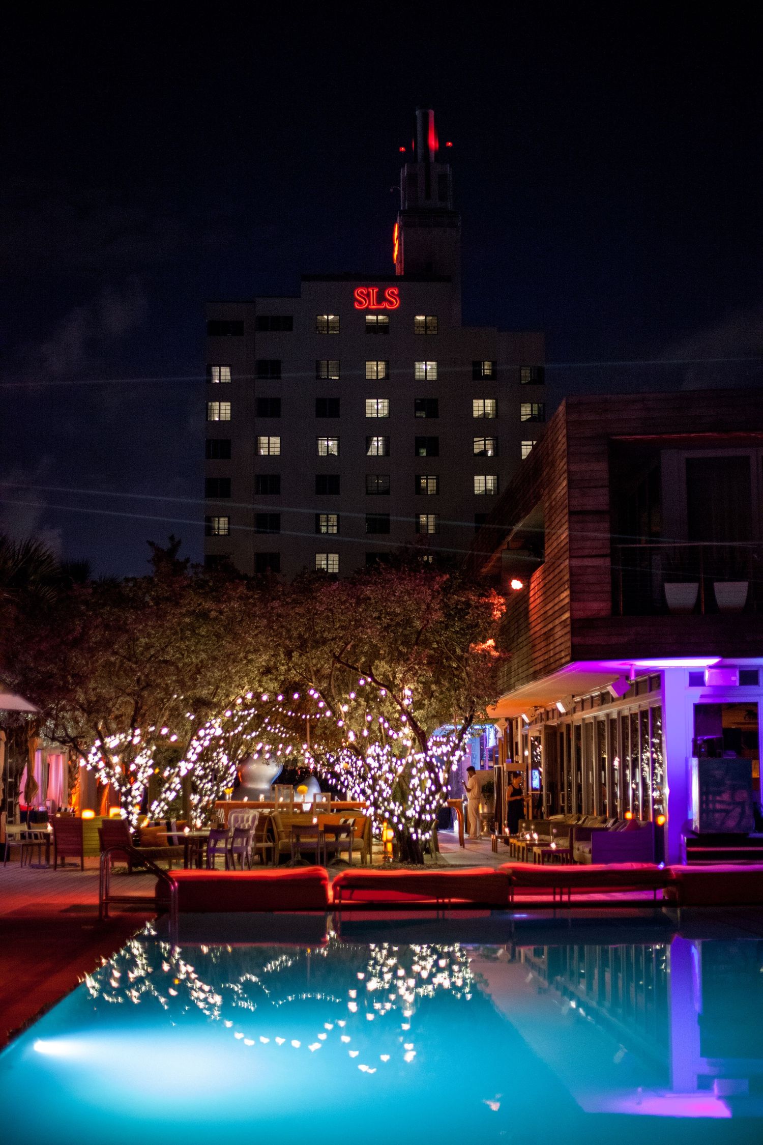 Illuminated hotel pool at night with decorative trees and purple accent lighting.