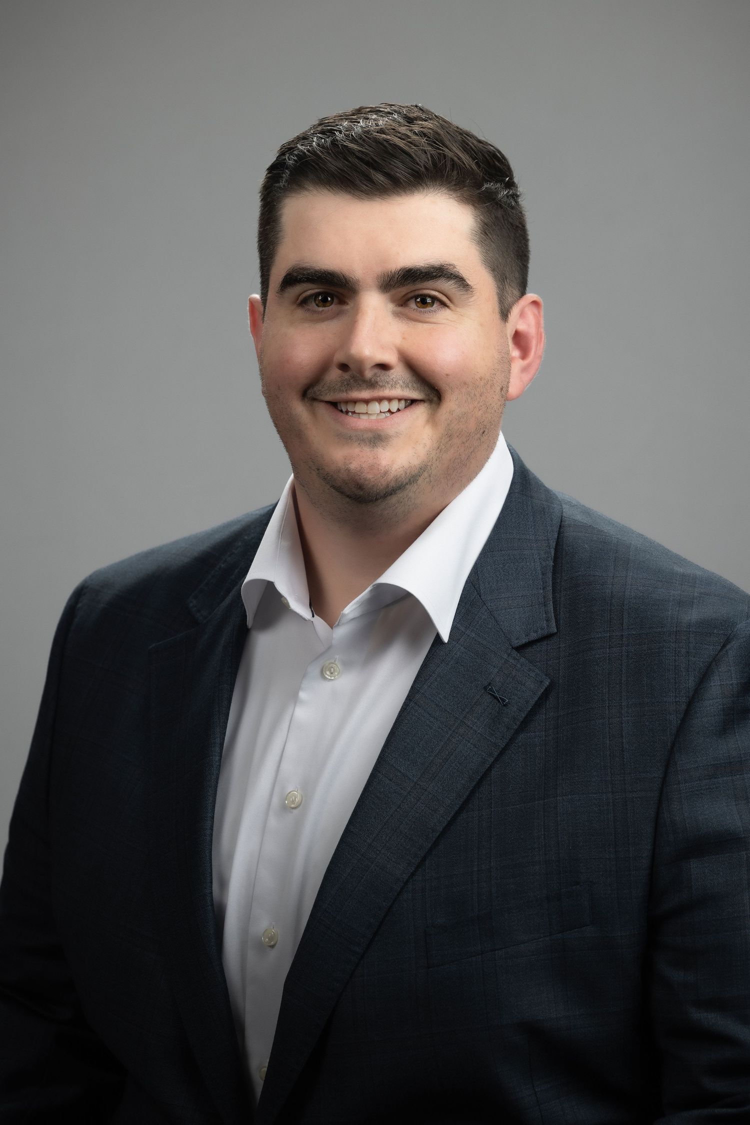 Professional headshot of a person wearing a dark suit jacket and white dress shirt against a gray background.