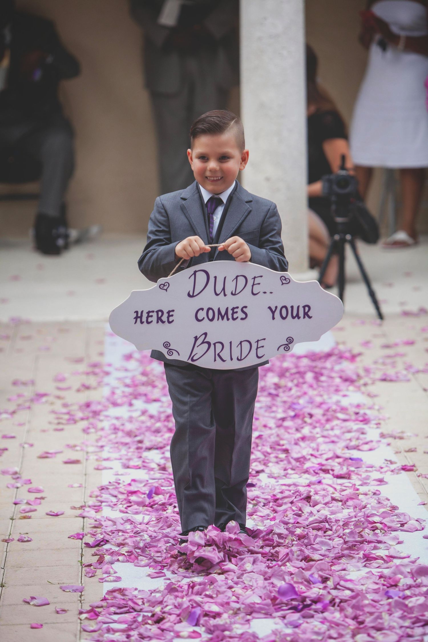 Young ring bearer holding sign on petal-covered walkway at wedding ceremony.
