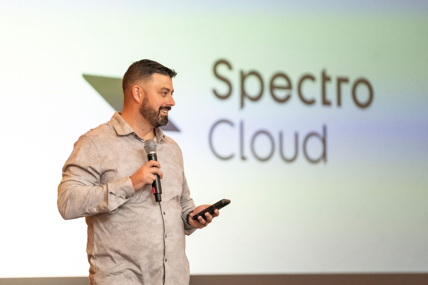 A speaker wearing a gray shirt holds a phone and microphone while presenting on stage with projected text behind.