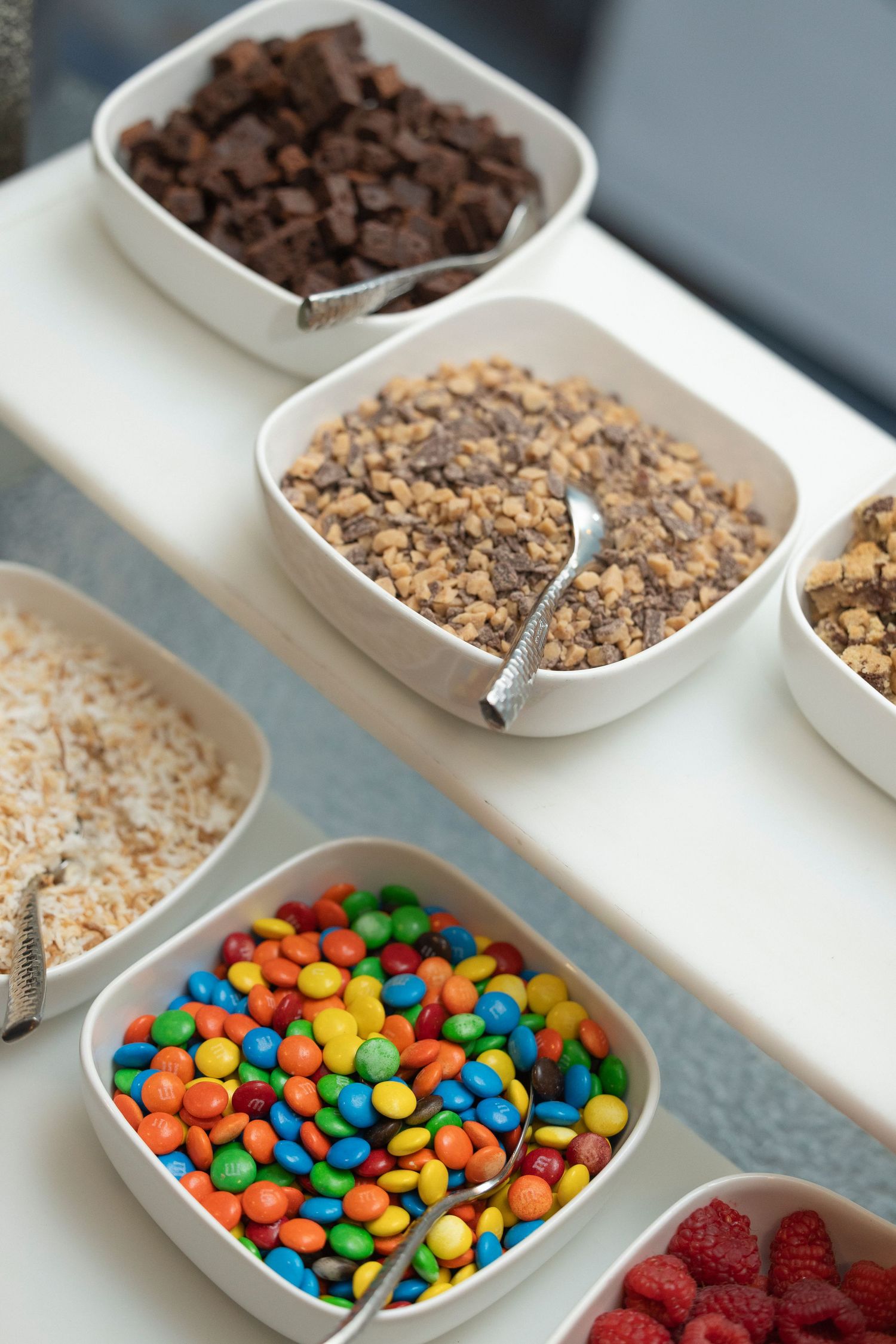 Colorful M&M candies, chocolate chips, and other toppings arranged in small white serving bowls for dessert preparation.