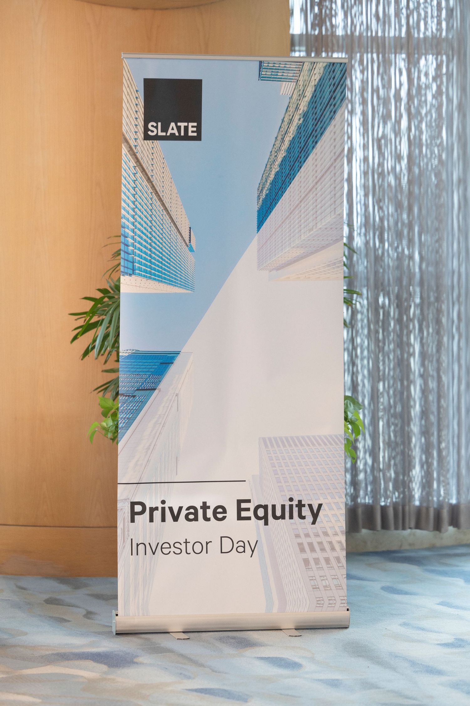 White banner displays 'Private Equity Investor Day' text against light blue wall with sheer curtains and potted plants.