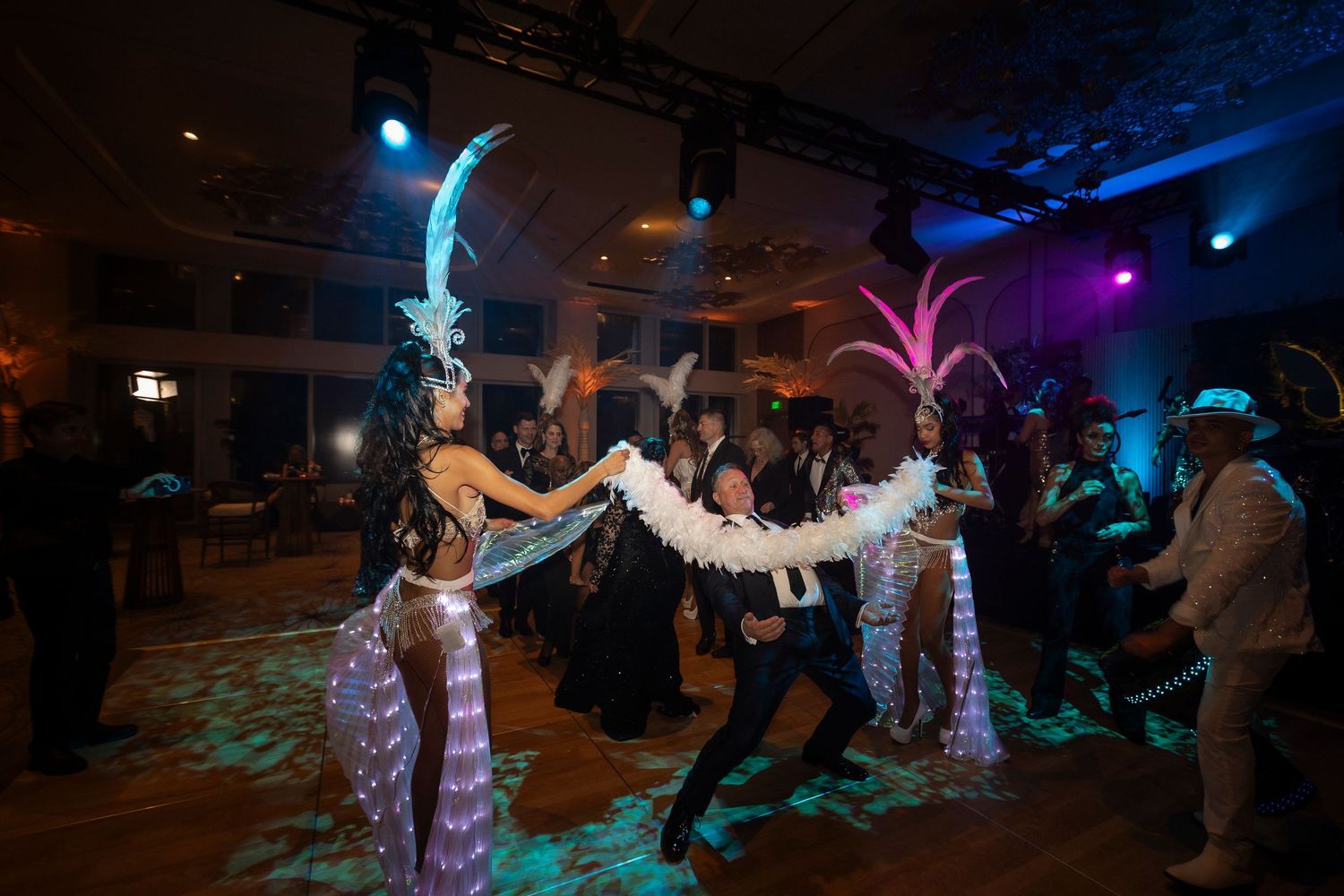 Dancers in vibrant costumes perform at a club with colorful neon lights and feathered props on a dark dancefloor.