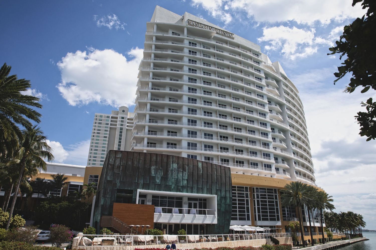 Modern curving high-rise condominium building with balconies and palm trees against a bright blue sky.