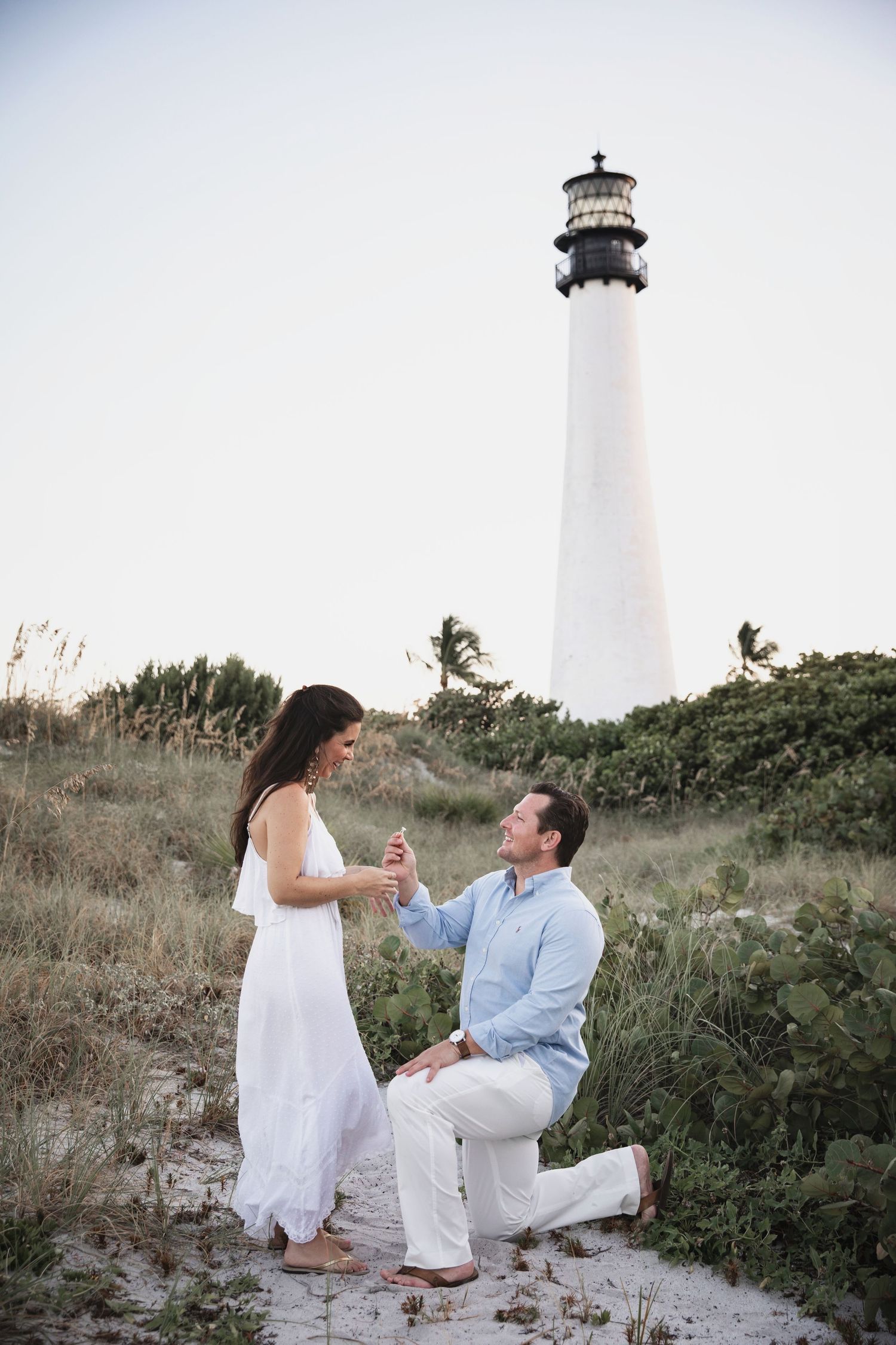 A proposal scene unfolds near a majestic white lighthouse on a rocky coastal setting.